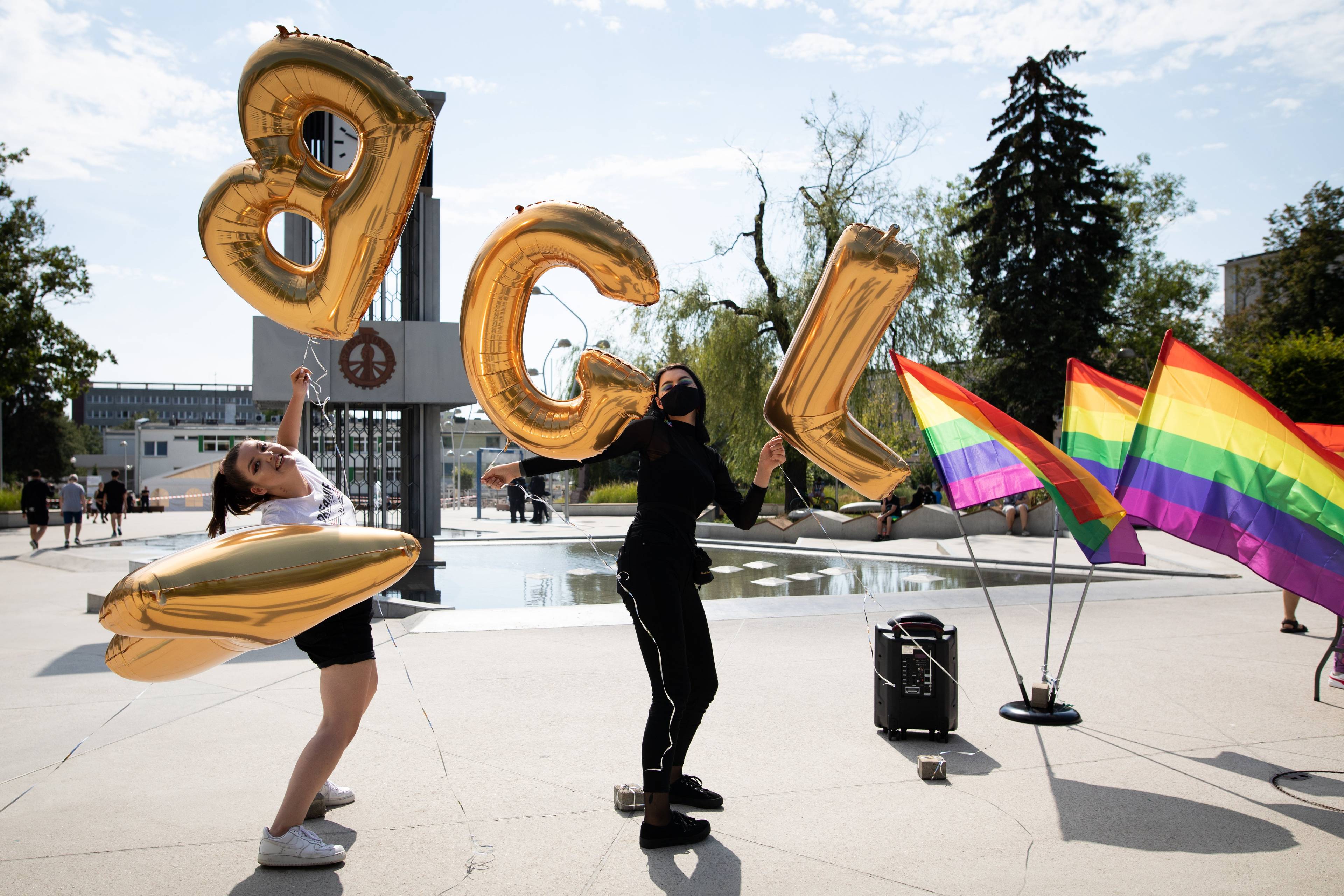 Dwie osoby trzymają balony w kształcie liter LGBT na placu miejskim, w tle powiewają tęczowe flagi