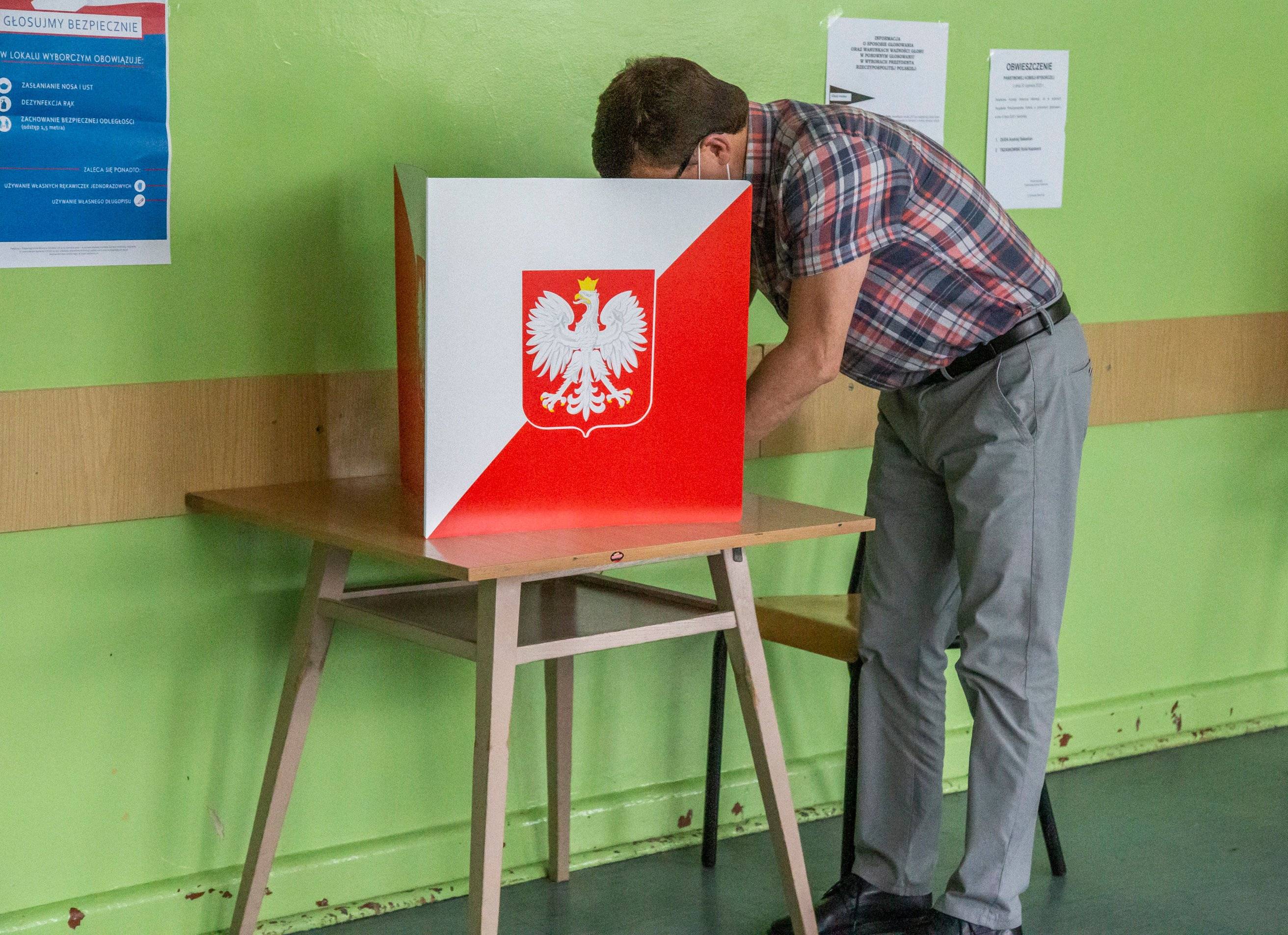 na zdjęciu mężczyzna pochyla się nad stolikiem osłoniętym kartonem z godłem Polski, żeby zaznaczyć głos na karcie wyborczej
