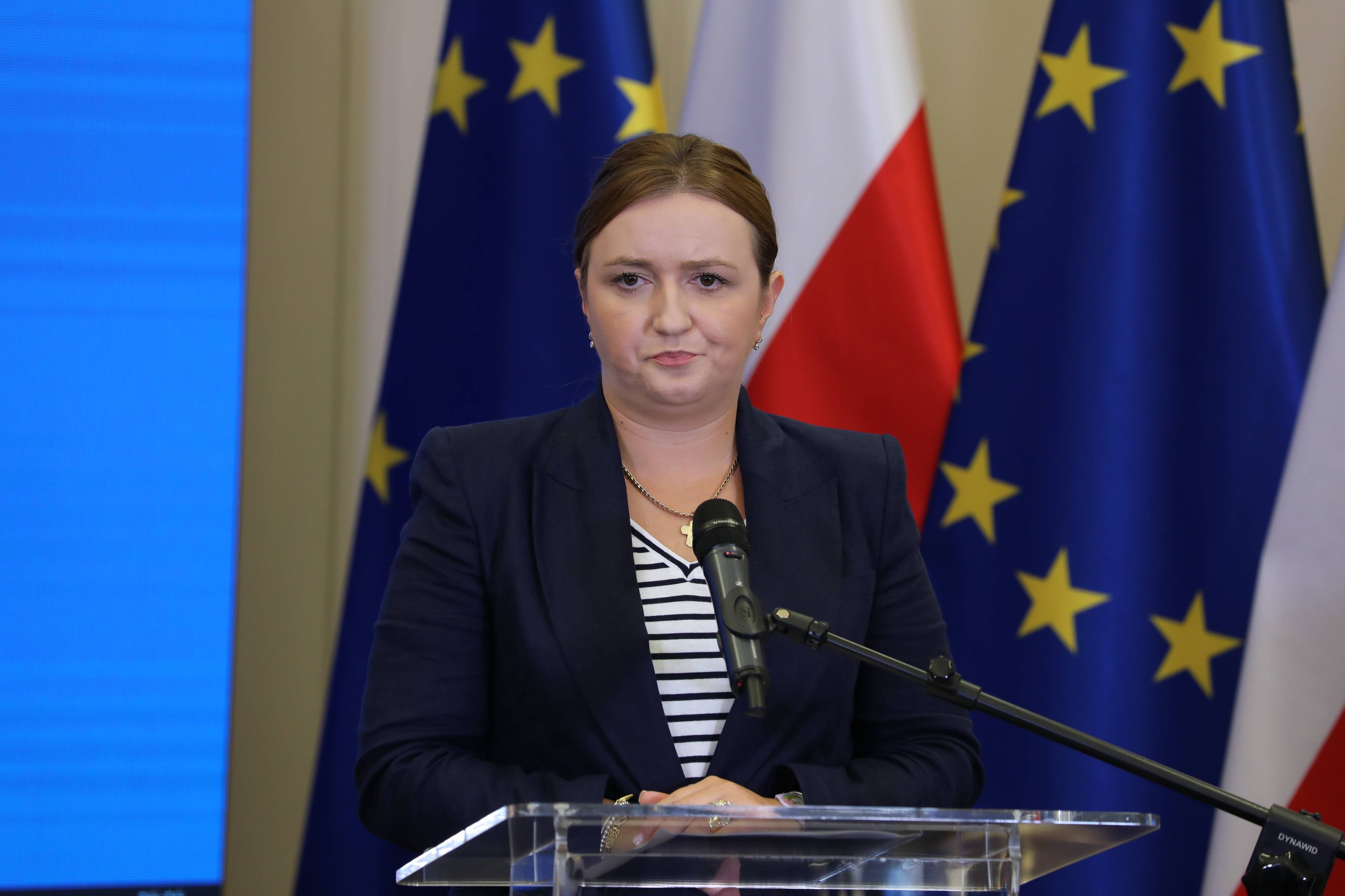 Kobieta na tle flag UE i PolskiOlga Semeniuk-Patkowska podczas konferencji prasowej na tle flag Polski i Unii Europejskiej