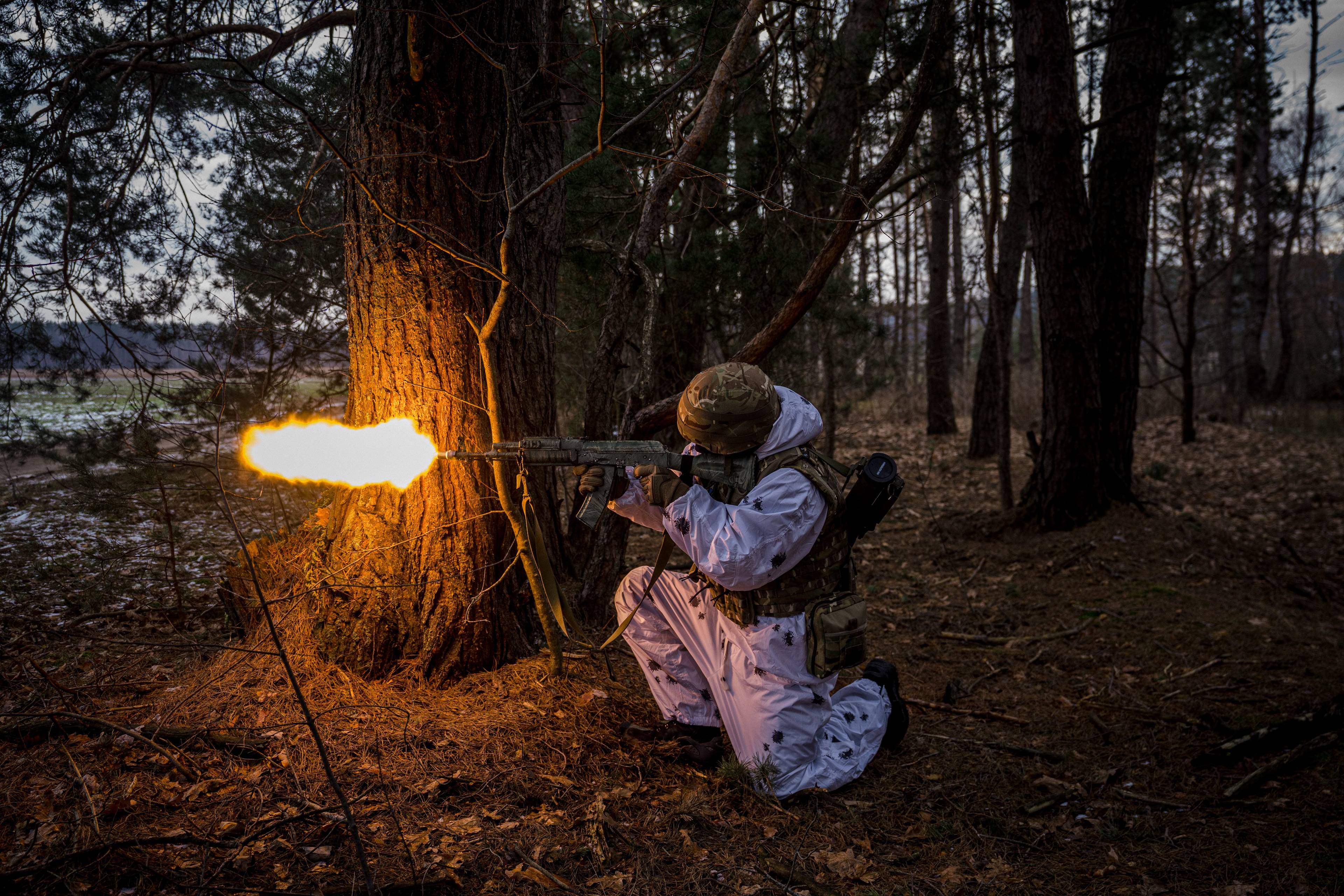 Żołnierz w białym uniformie klęka i strzela z broni maszynowej na granicy sosnowego lasu