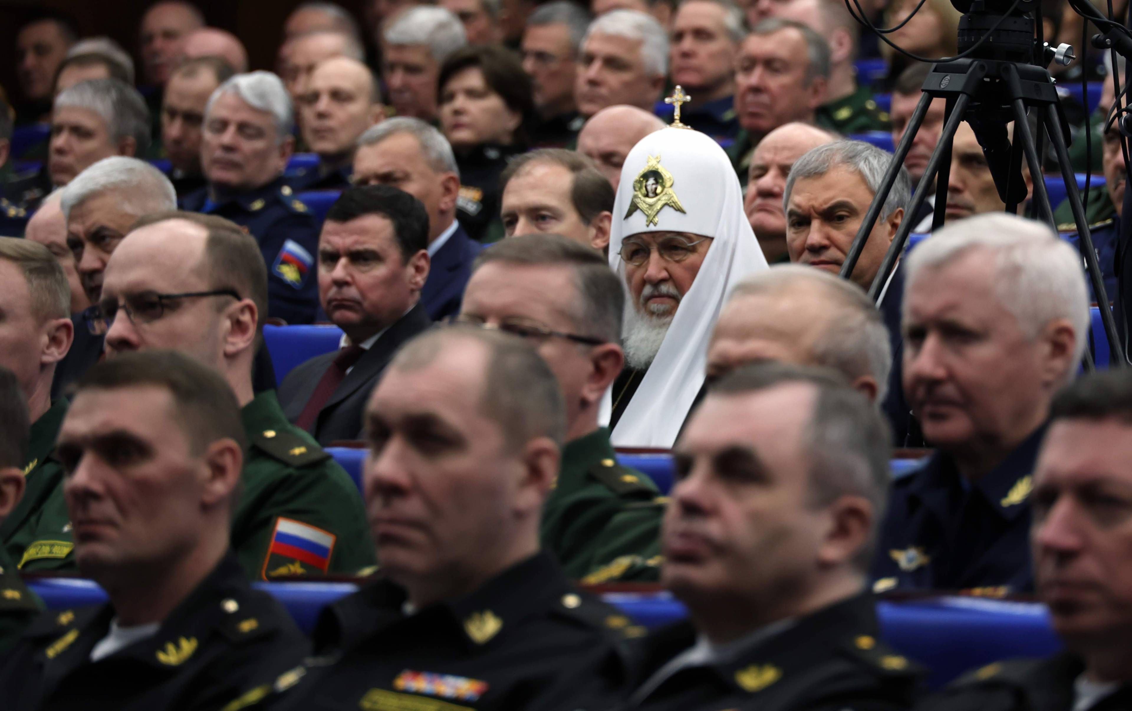 duchowny prawosławny a ceremonialnym białym nakryciu głowy z krzyżem siedzi na sali wśród wojskowych