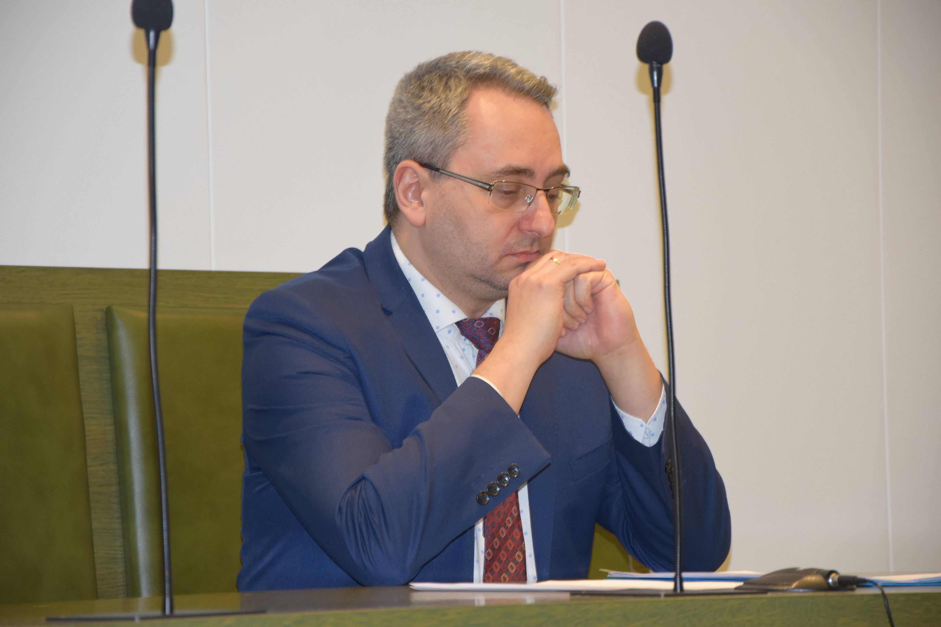 Mężczyzna w okularach siedzi za stołme w sali sądowej. Michał Lasota
