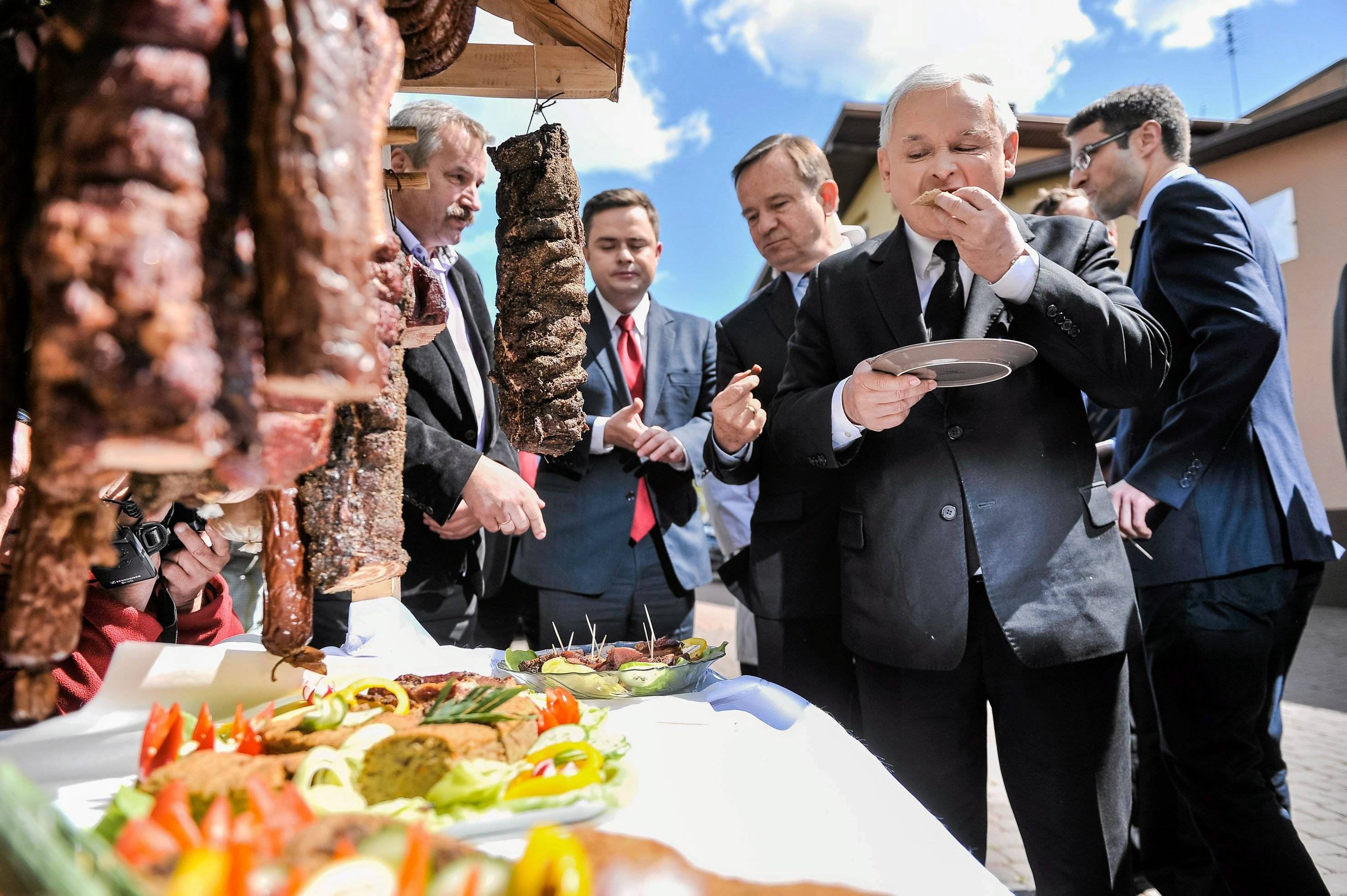 Jarosław Kaczyński stoi obok stoiska z mięsami, zjadając coś z talerzyka
