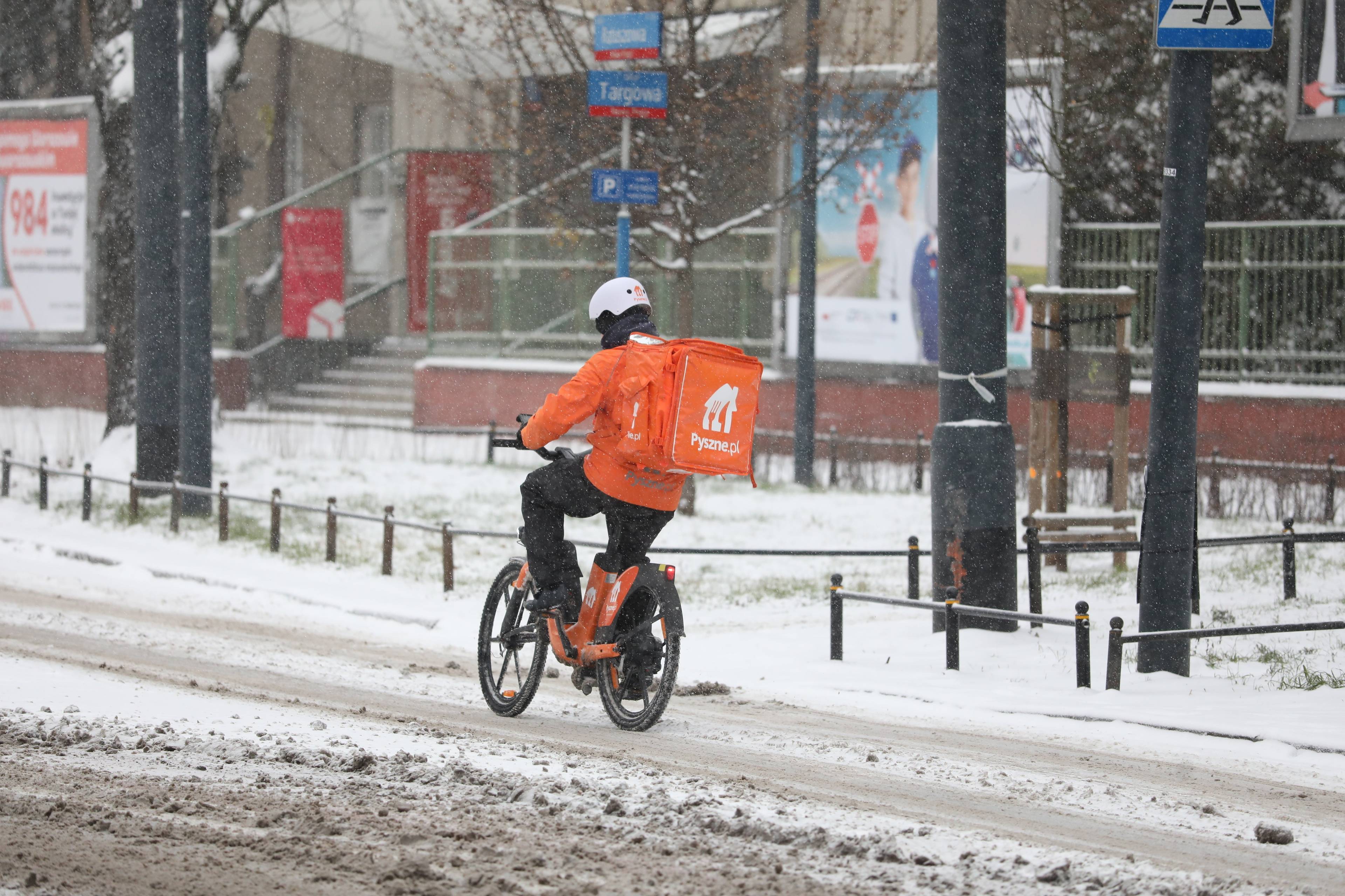 Kurier z plecakiem pyszne.pl jedzie rowerem po zaśnieżonej ulicy