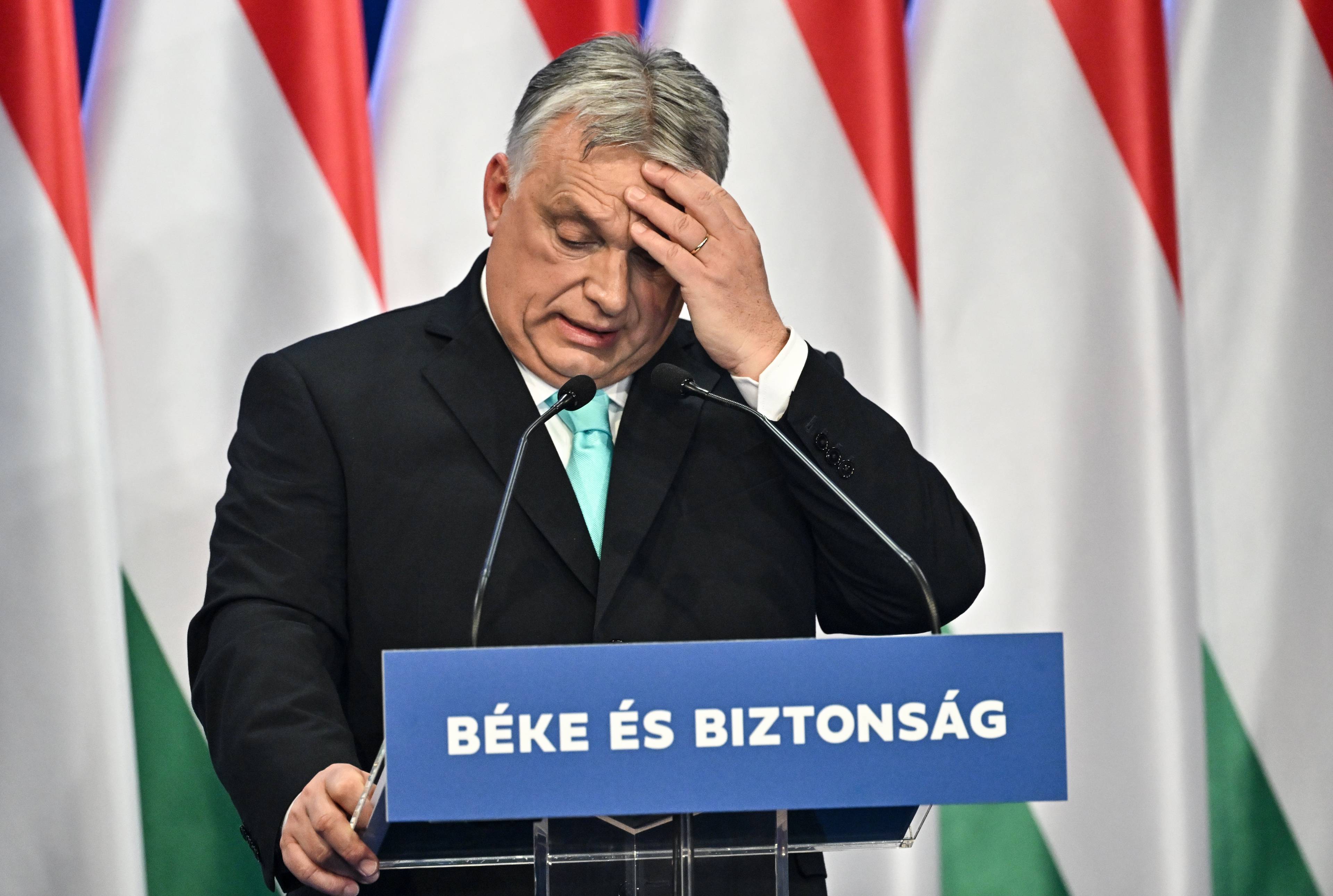 Mężczyzna w wieku ok 60 lat stoi na mównicy i chwyta się za czoło. To Viktor Orban. W tle flagi Węgier