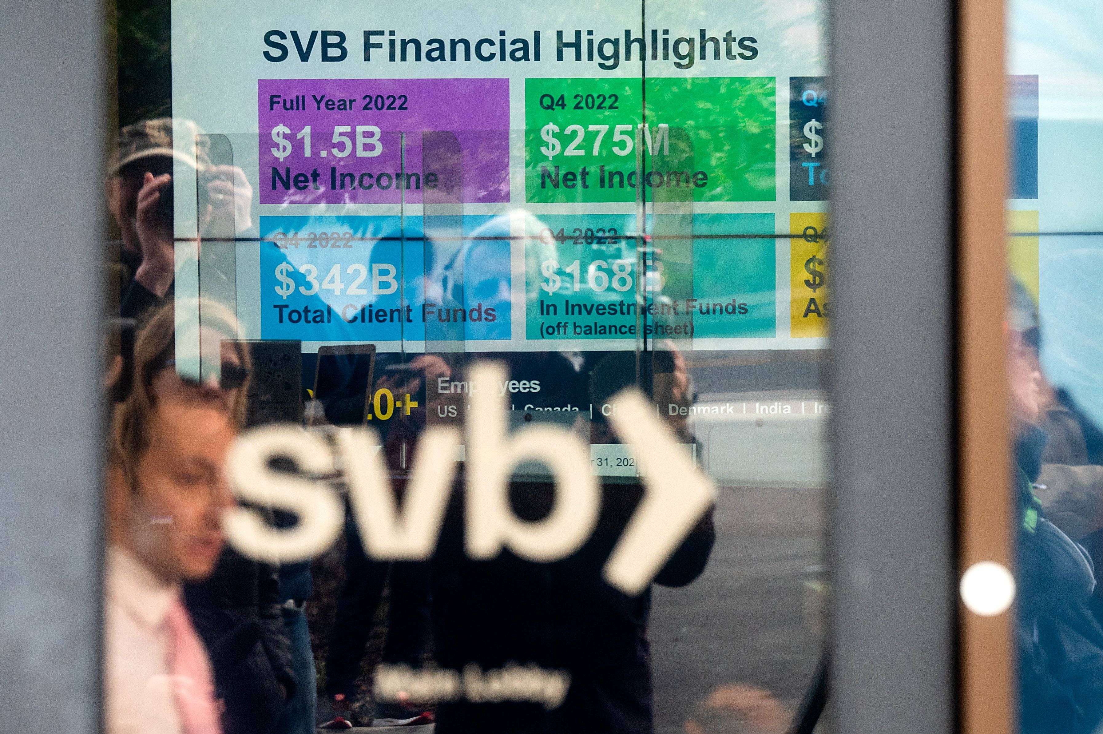 Na pierwszym planie szyba, za nią wyświetlacz wymienia osiągnięcia Silicon Valley Bank (SVB), gdy klienci zbierają się, aby wypłacić pieniądze