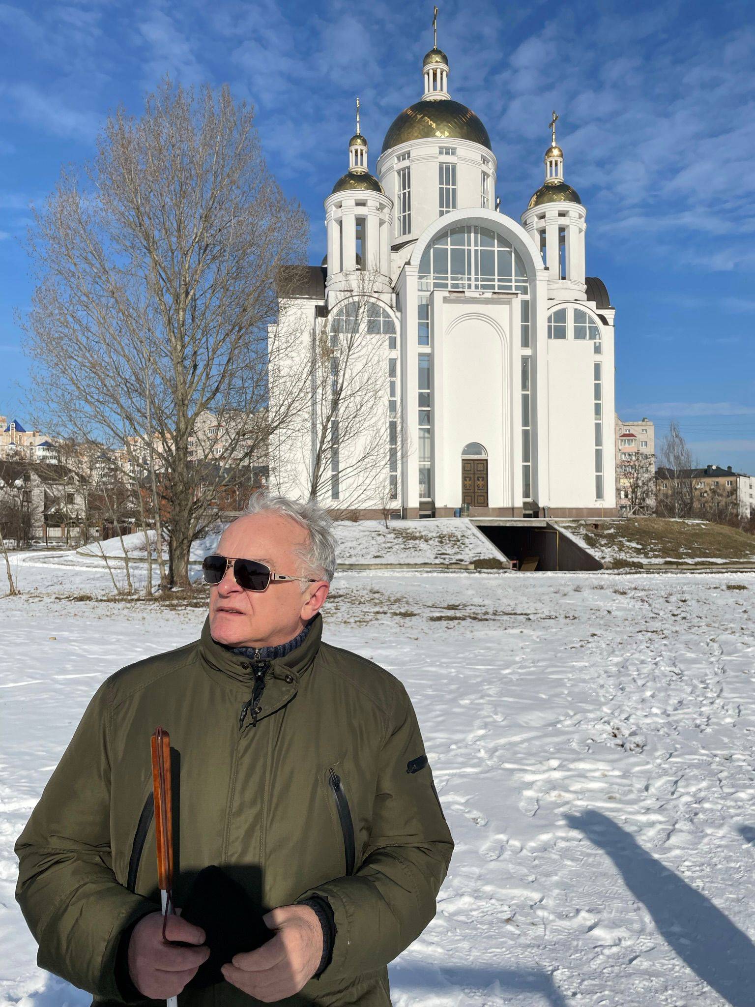 człowiek w ciemnych okularach trzymający białą laskę, w tle biała cerkiew