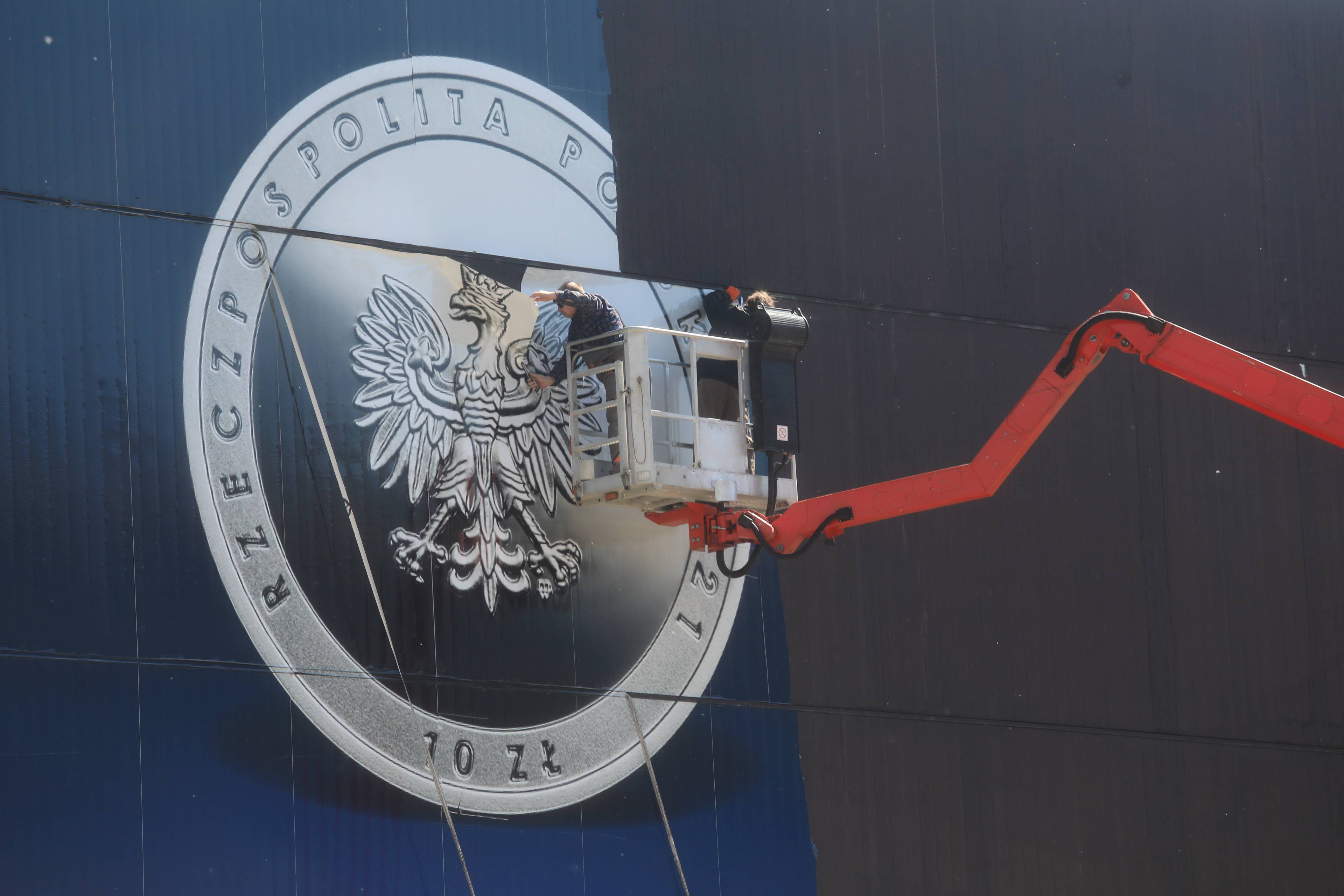 Robotnicy na podnośniku demontują baner z elewacji Narodowego Banku Polskiego. Na banerze polska moneta