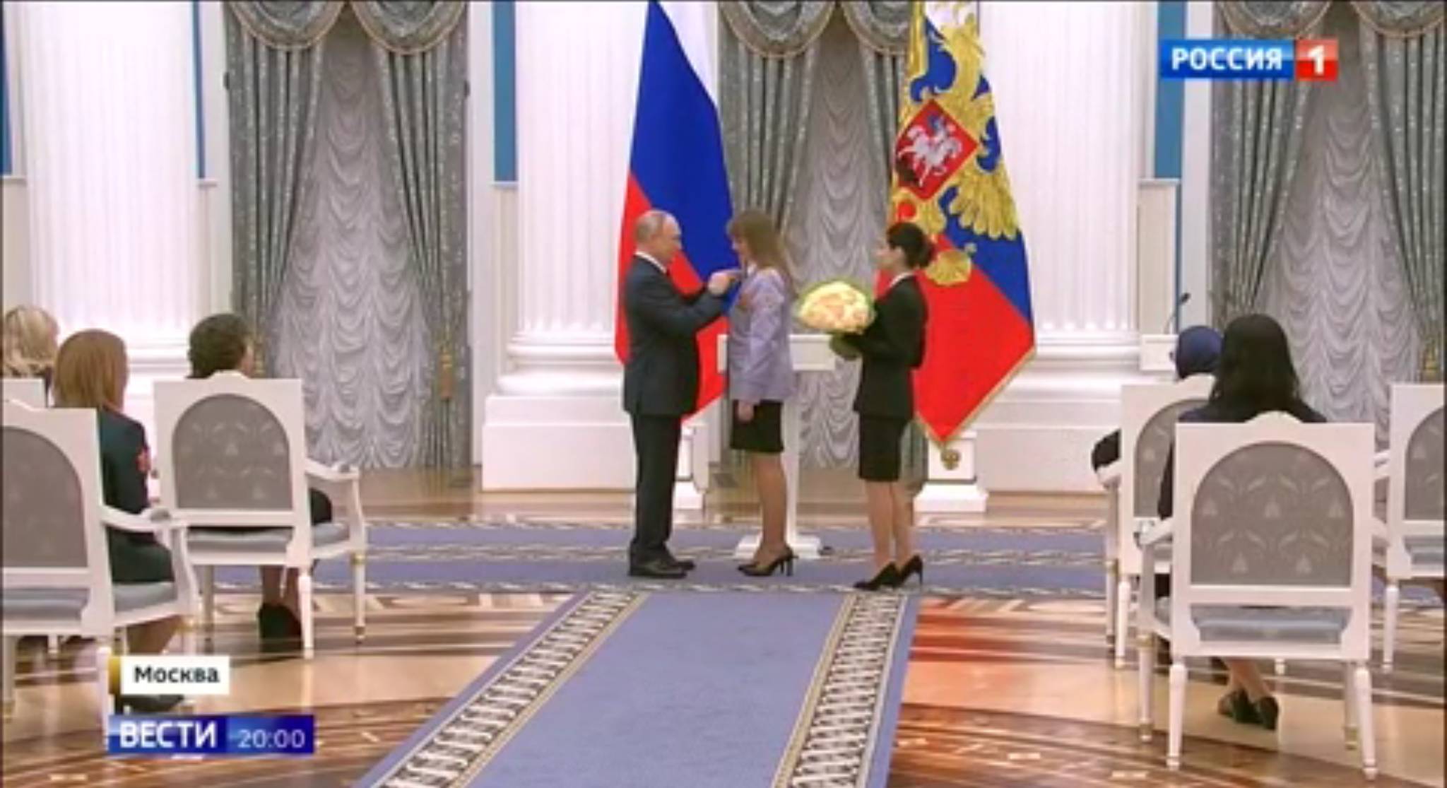 Putin w sali pałacowej przypina kobiecie order. Z boku stoi inna kobieta z bukietem żołtych kwiatów, które Putin zaraz wręczy odznaczonej