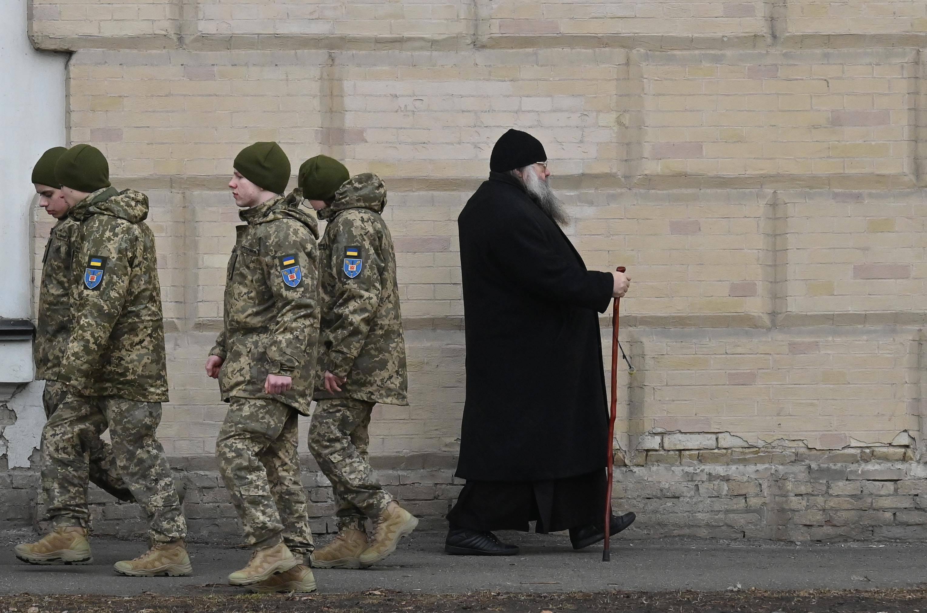Prawosławny duchowny mija grupę ukraińskich żołnierzy na tle muru. Ukraińska Cerkiew Prawosławna.