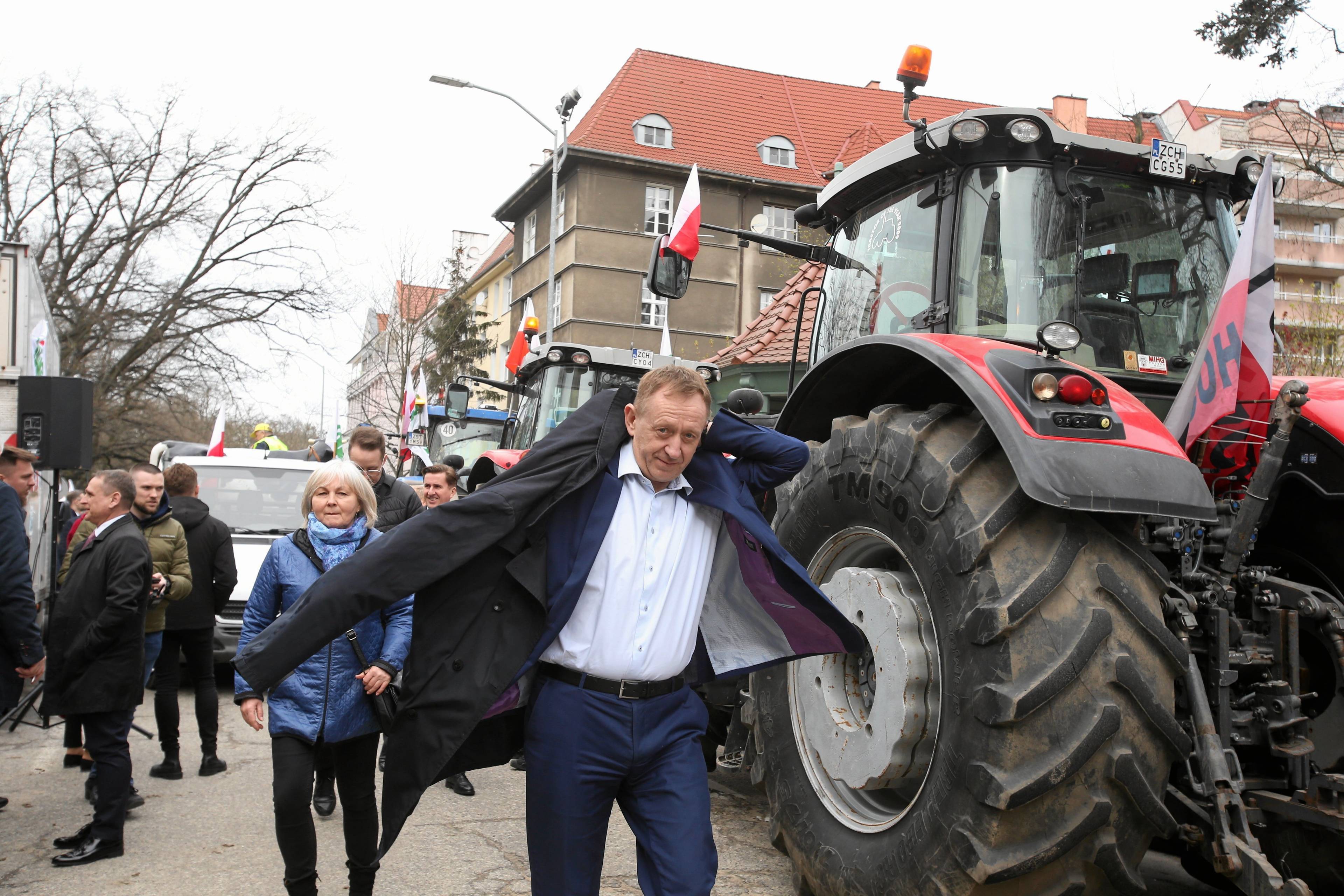 Minister Robert Telus po rozmowach z protestującymi rolnikami ubiera płaszcz i oddala się od rozmówców. W tle ciągnik rolniczy.