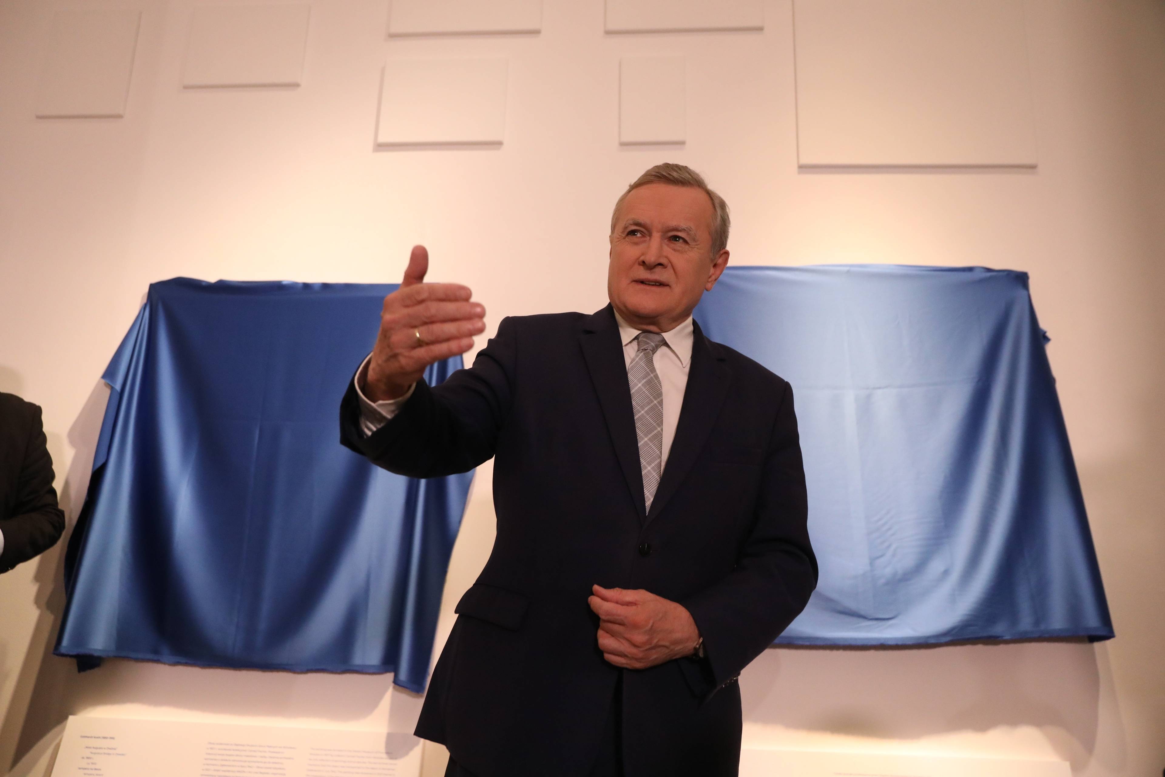 Minister Kultury Piotr Gliński w garniturze wskazuje na coś ręką, za nim przykryte niebieskimi narzutami ekspozycja w galerii