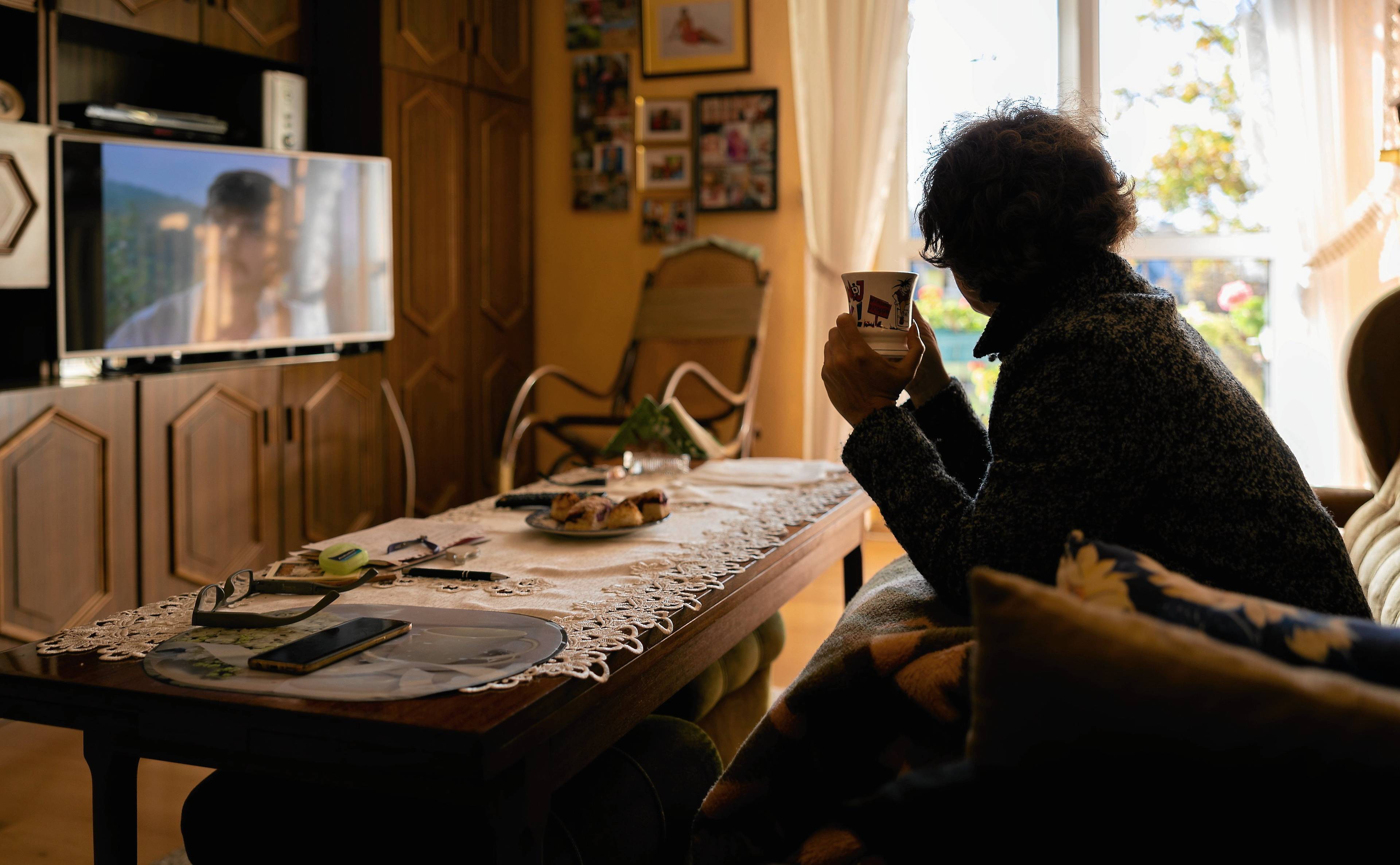 Kobieta siedzi na kanapie w mieszkaniu, jest przykryta kocem w dłoniach trzyma kubek z herbatą