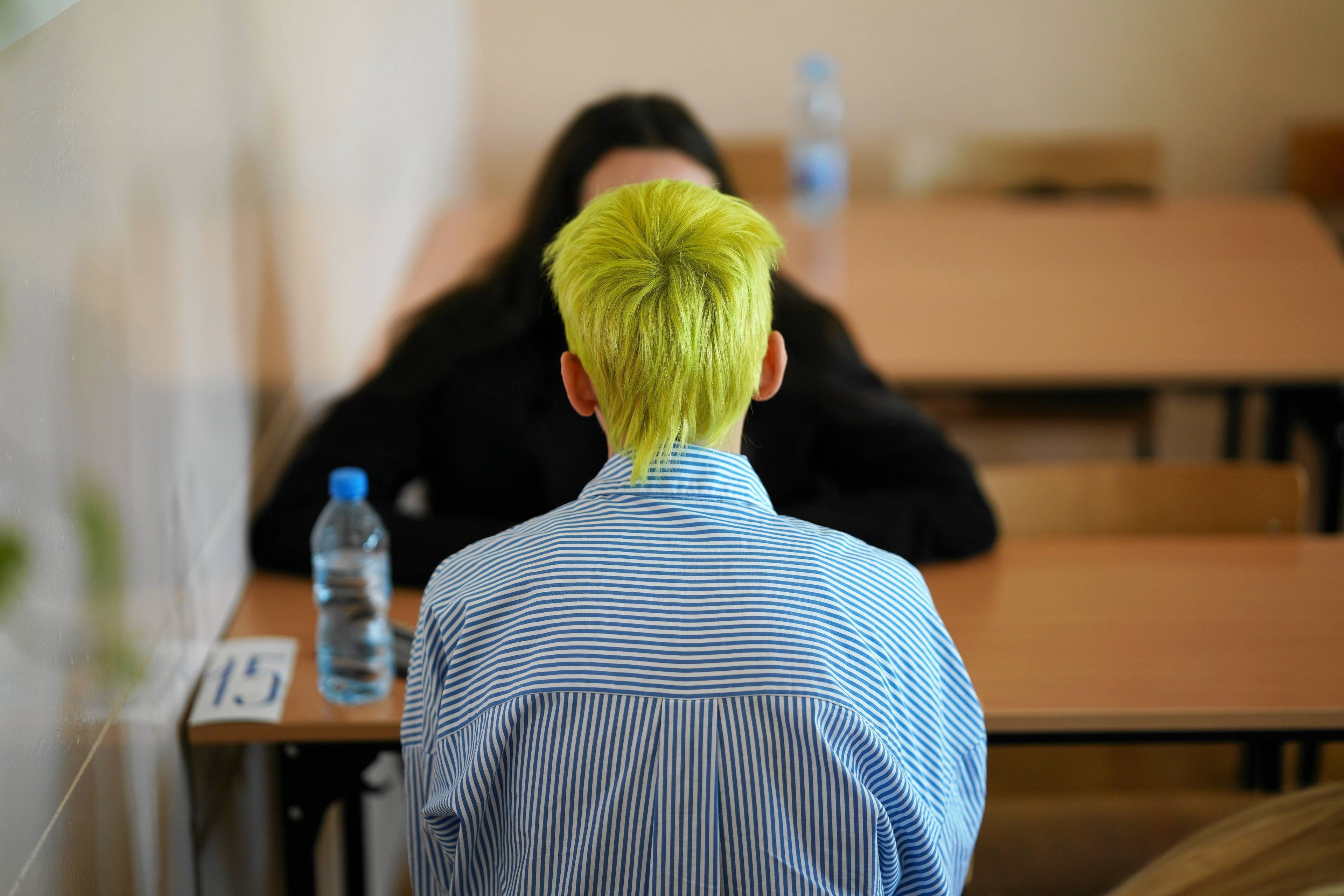 Na zdjęciu osoba w farbowanych żółtych włosach siedzi tyłem do obiektywu przy szkolnej ławce. Chwila przed maturą z języka polskiego