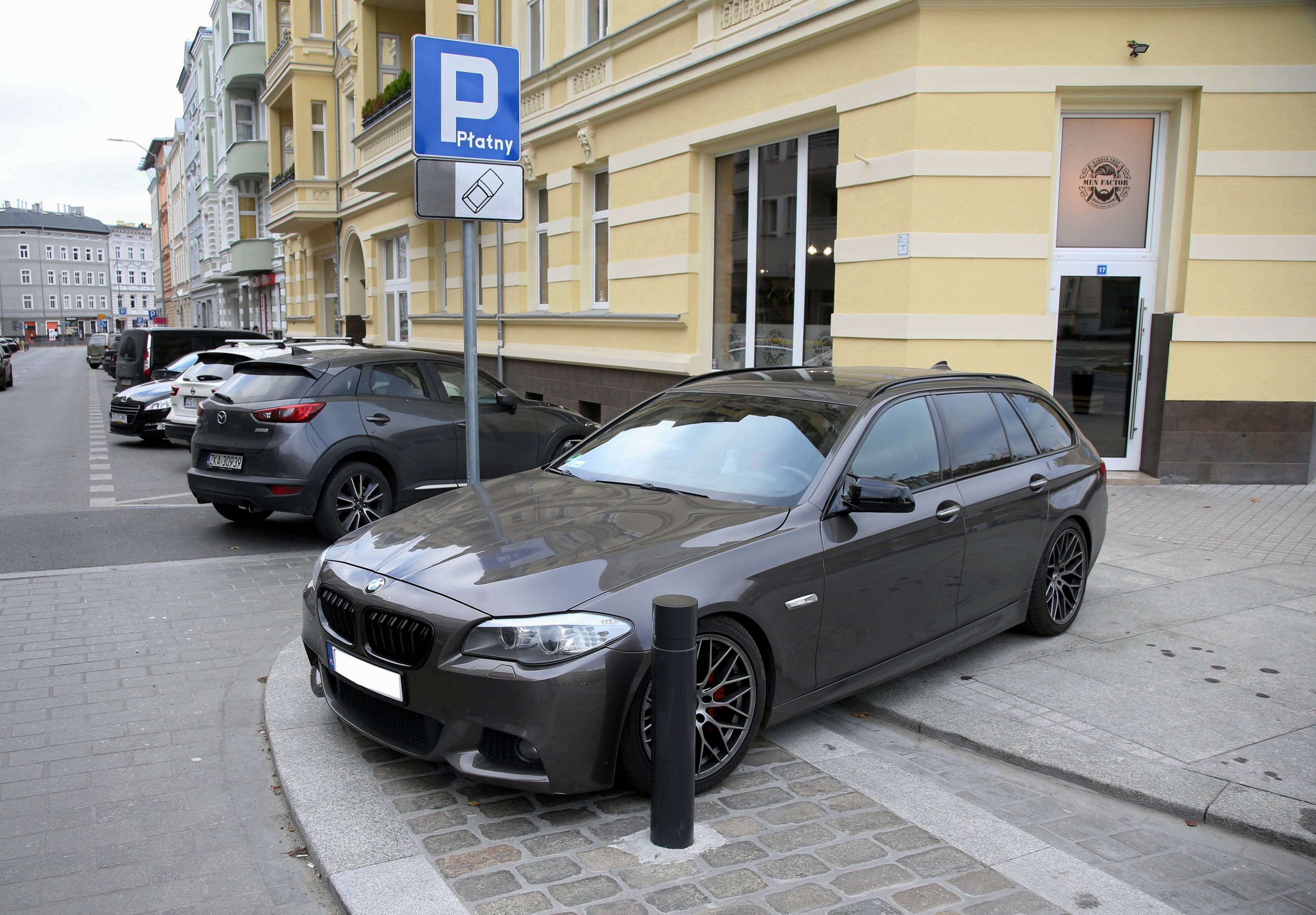 Czarne BMW parkujące nieprawidłowo i zajmujące cały chodnik przy słupku, który powinien utrudnić postawienie tam samochodu