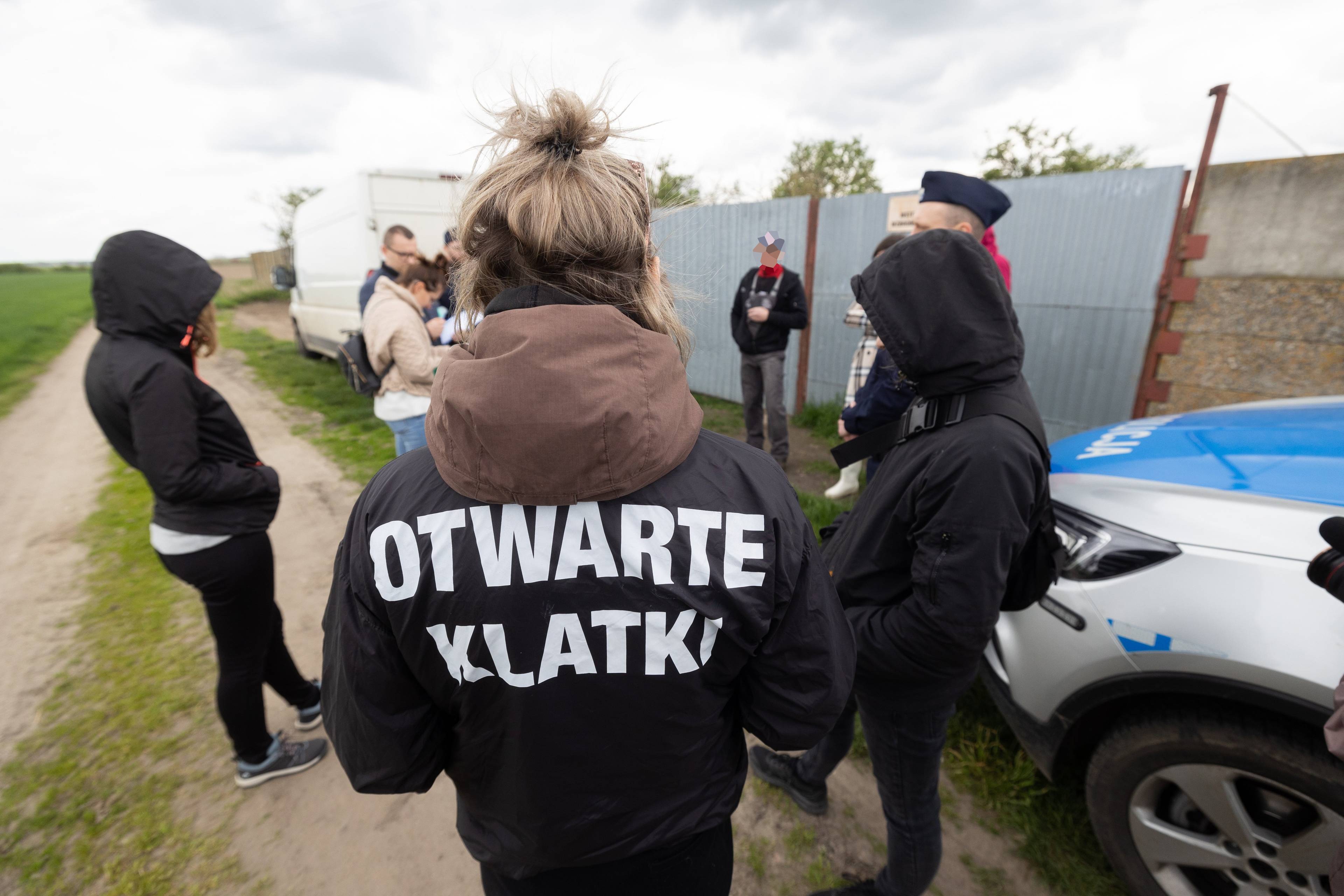 Kobieta w kurtce z napisem otwarte klatki stoi w towarzystwie policji przed bramą fermy