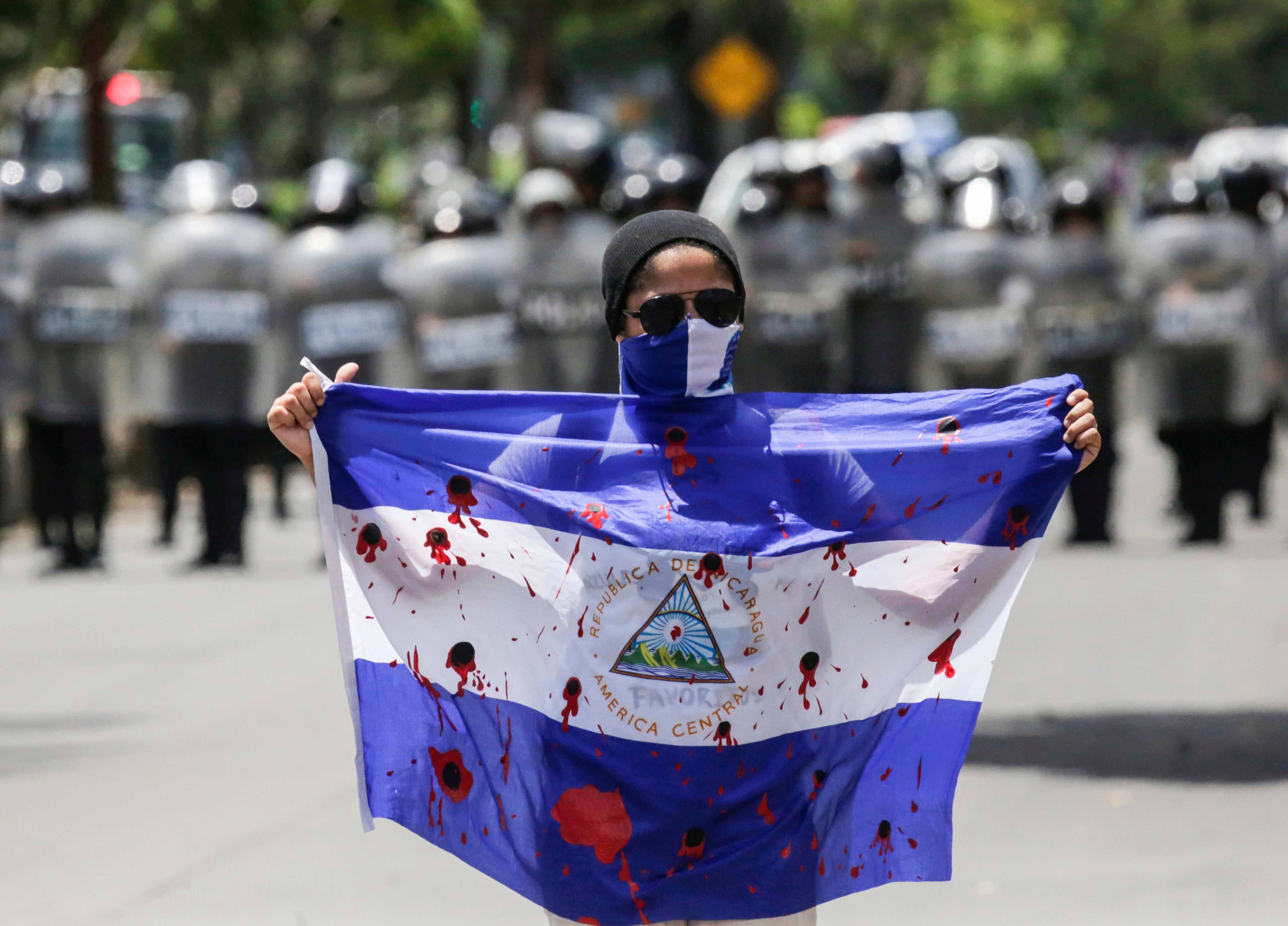 Demonstrant trzyma flagę niebiesko-biało-niebieską Nikaragui na której narysowano krwawe ślady od kul. W tle widać rząd policjantów z tarczami.