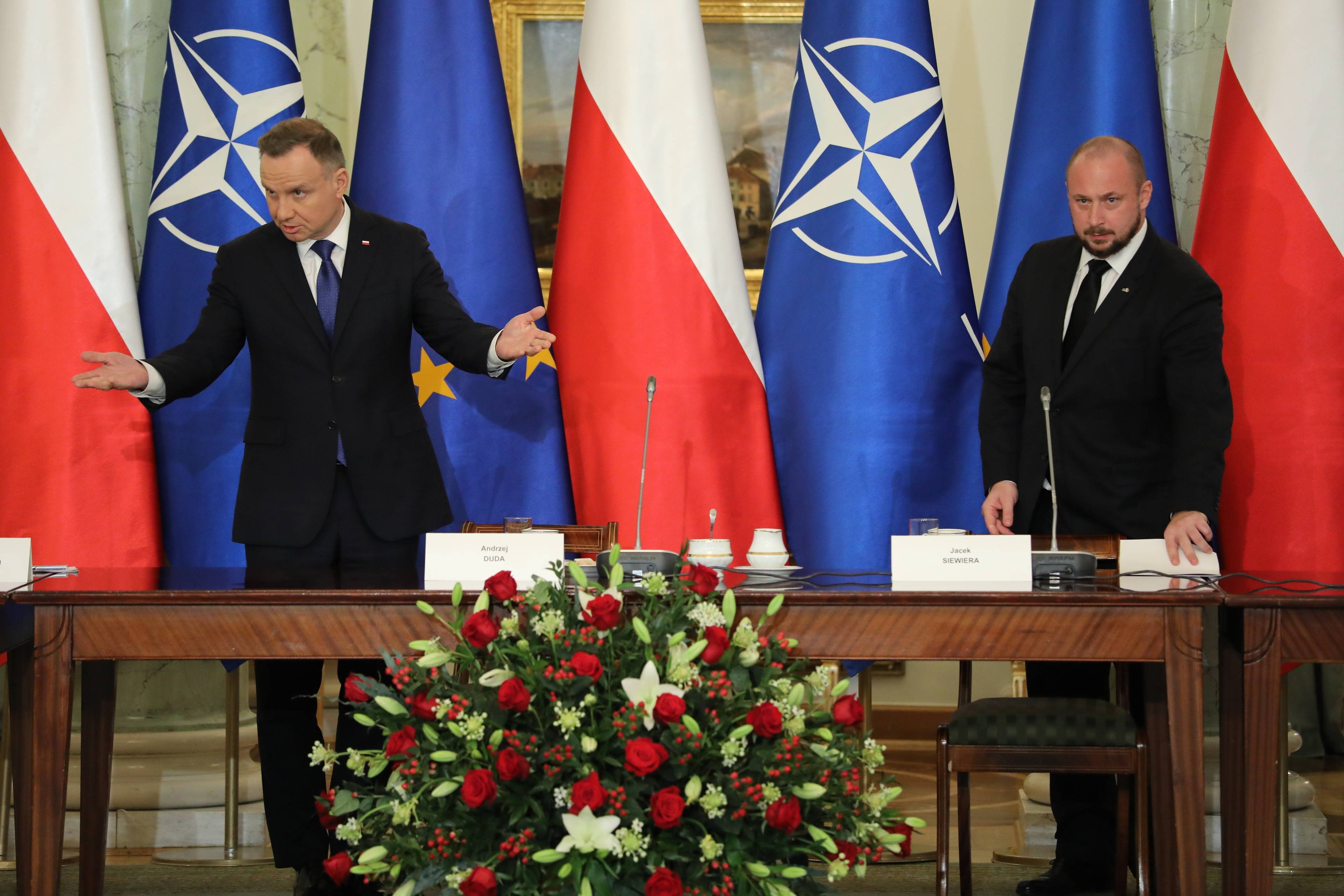 Dwaj mezczyźni w garniturach na tle flag Polski i NATO: Andrzej Duda i Jacek Siewiera. Duda rozkłada ręce/