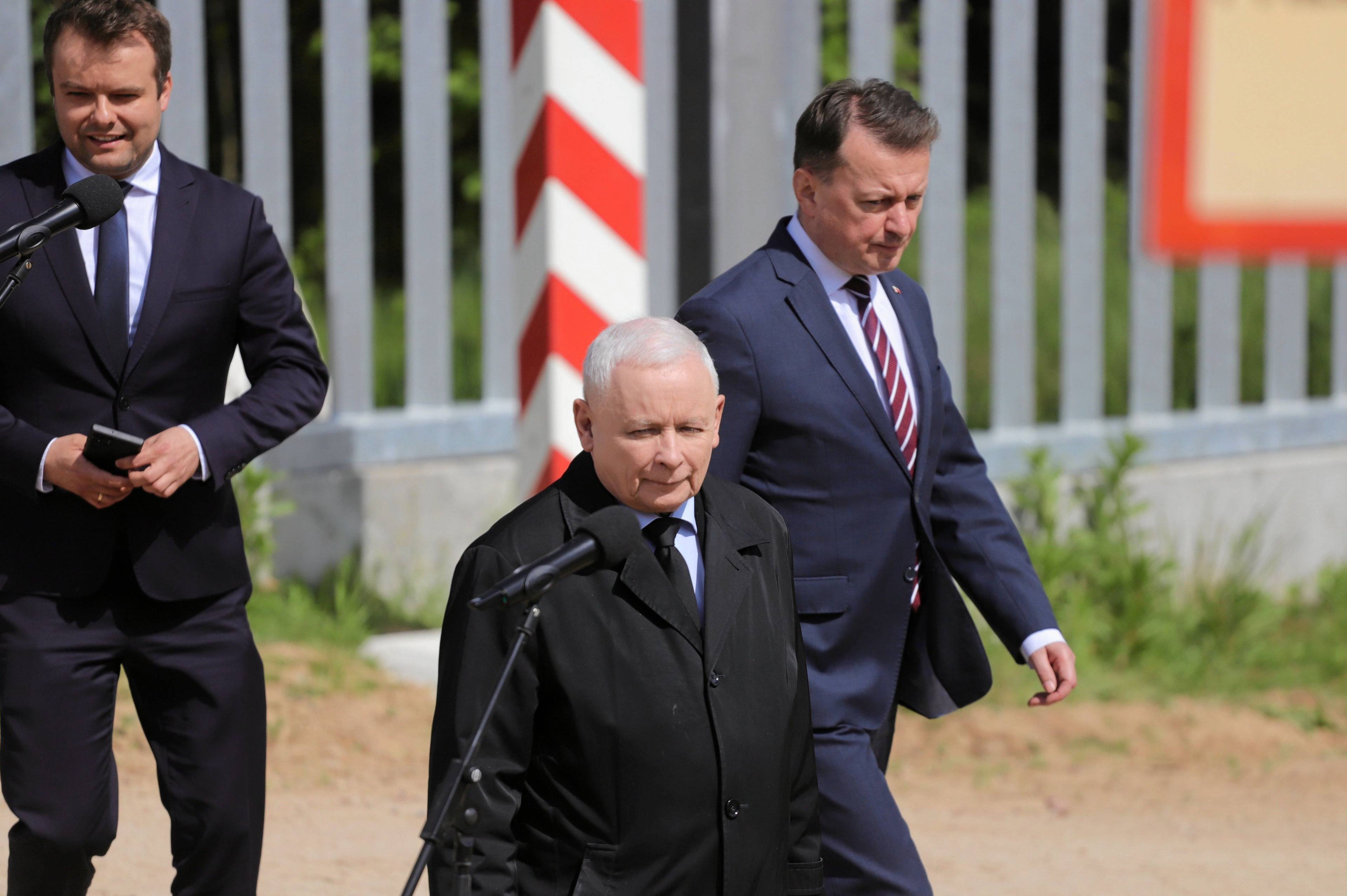 Rafał Bochenek, Jarosław Kaczyński i Mariusz Błaszczak stoją przed mikrofonami na tle płotu na granicy polsko-białoruskiej.