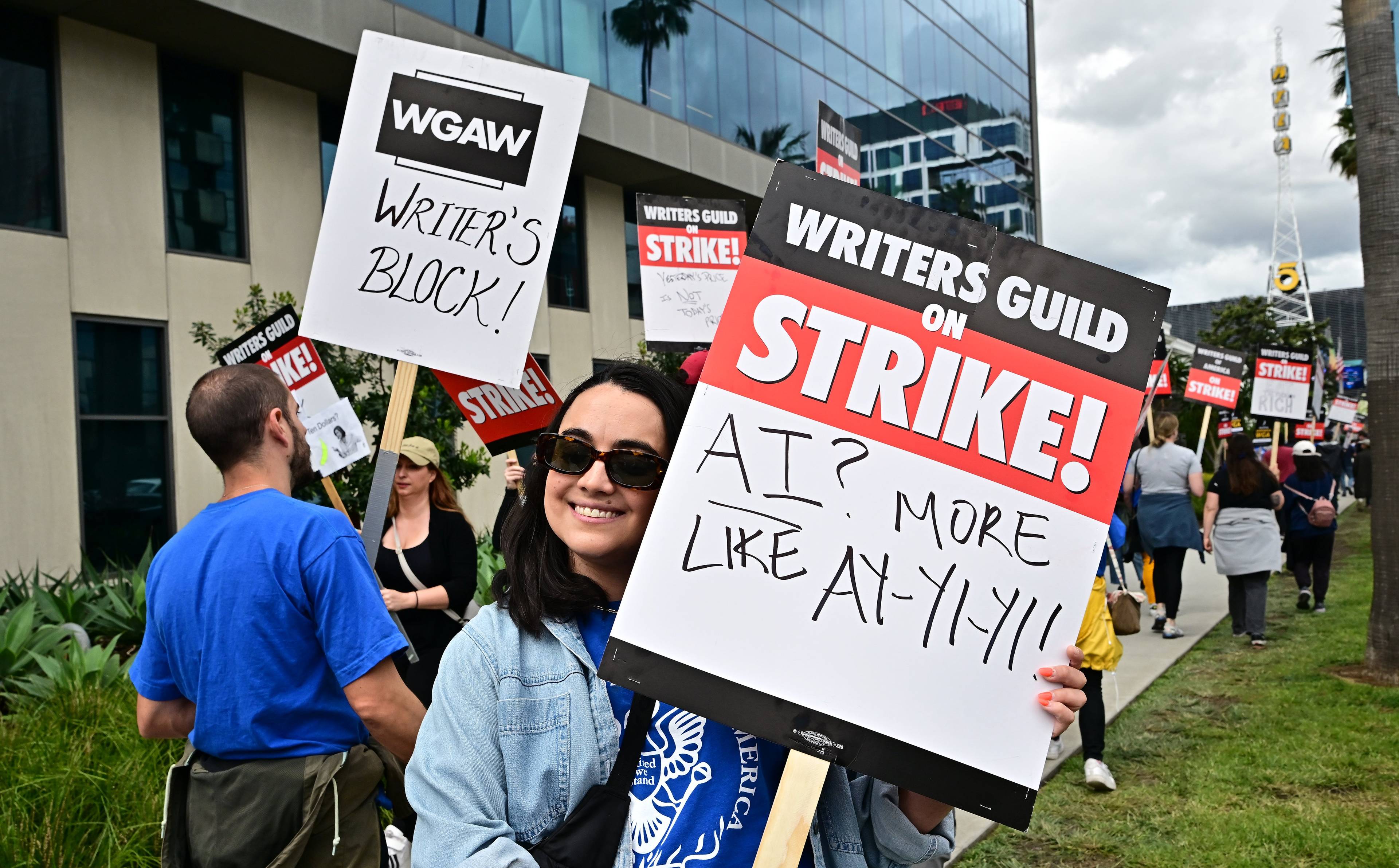 Kobieta stoi z tablicą, na której napisano po angielsku hasło „Writers Guild on Strike” (strajk związku scenarzystów), w tle inni demonstranci