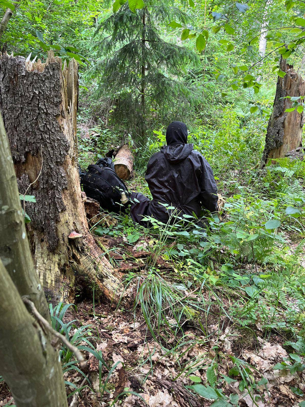 Mężczyzna w kapturze i płaszczu przeciwdeszczowym siedzi na trawie w środku lasu