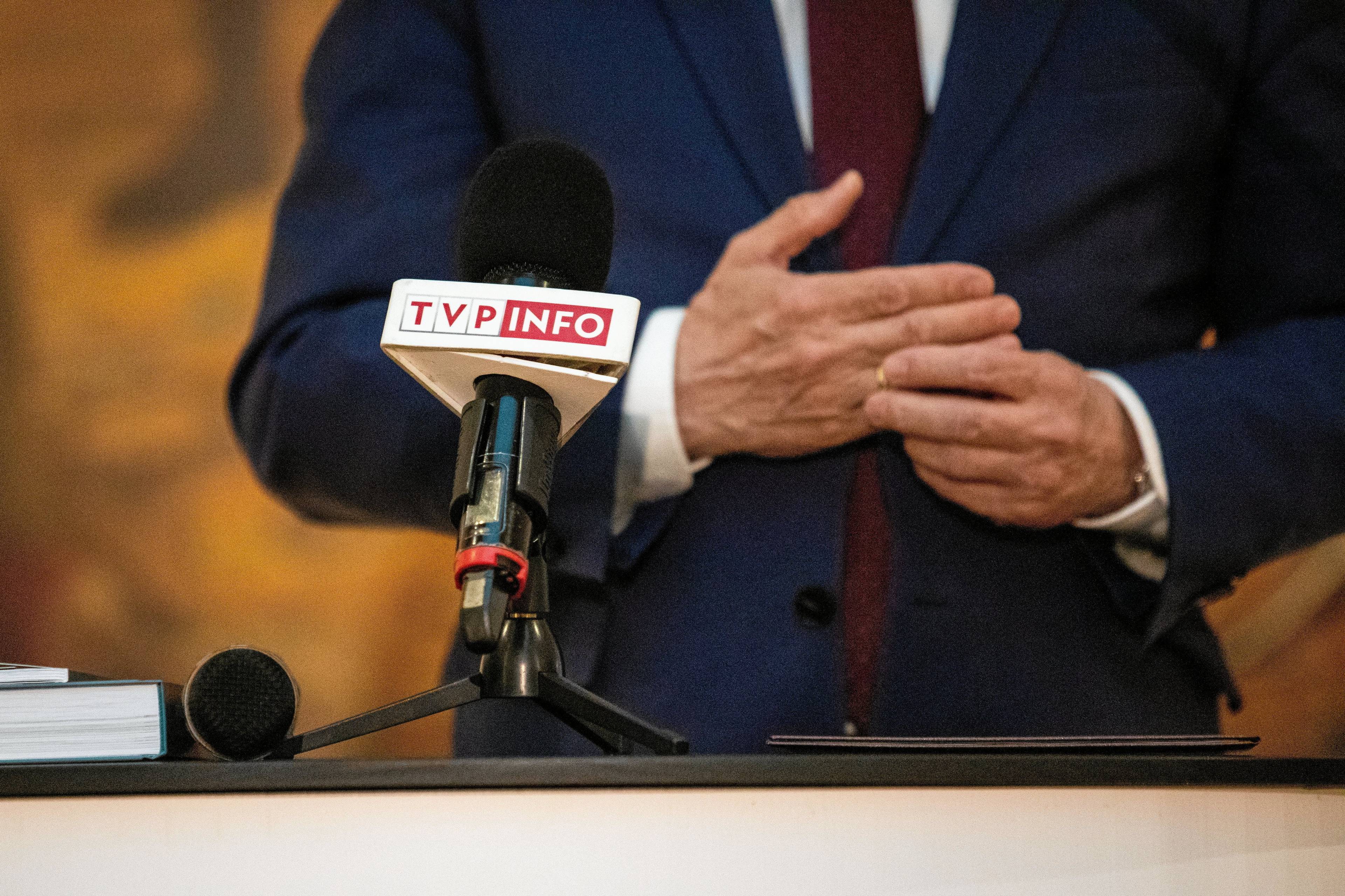 Mikrofon z napisem TVP Info, w tle dłonie mężczyzny w garniturze