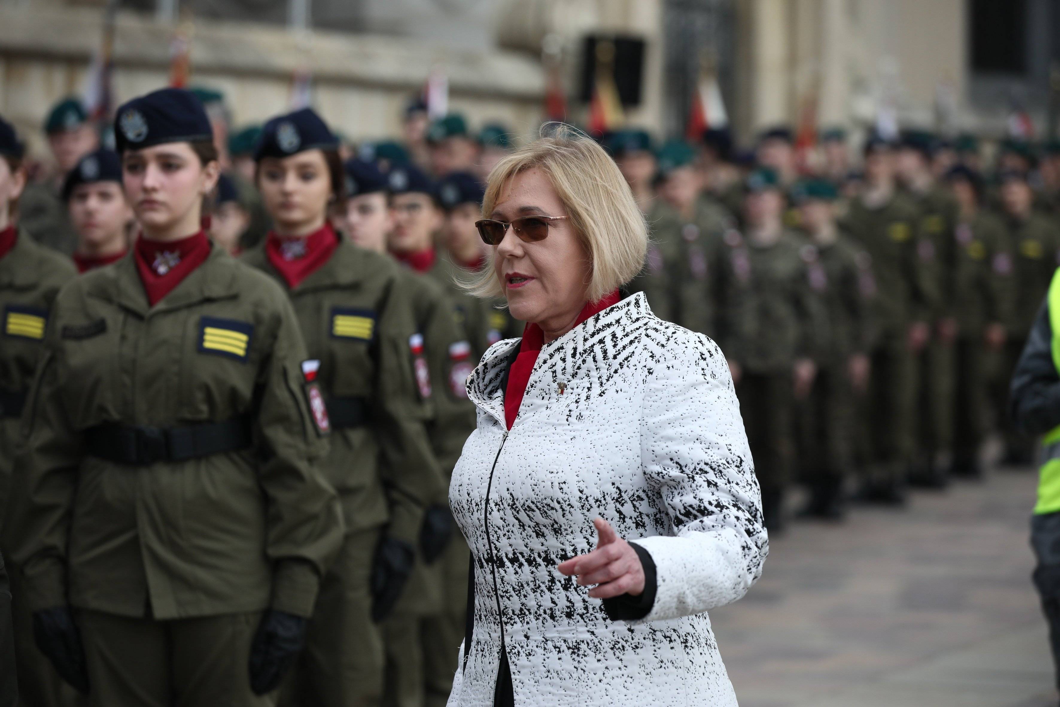 Kobieta w jasnej kurtce i ciemnych okularach stoi obok młodzieży w wojskowych mundurach