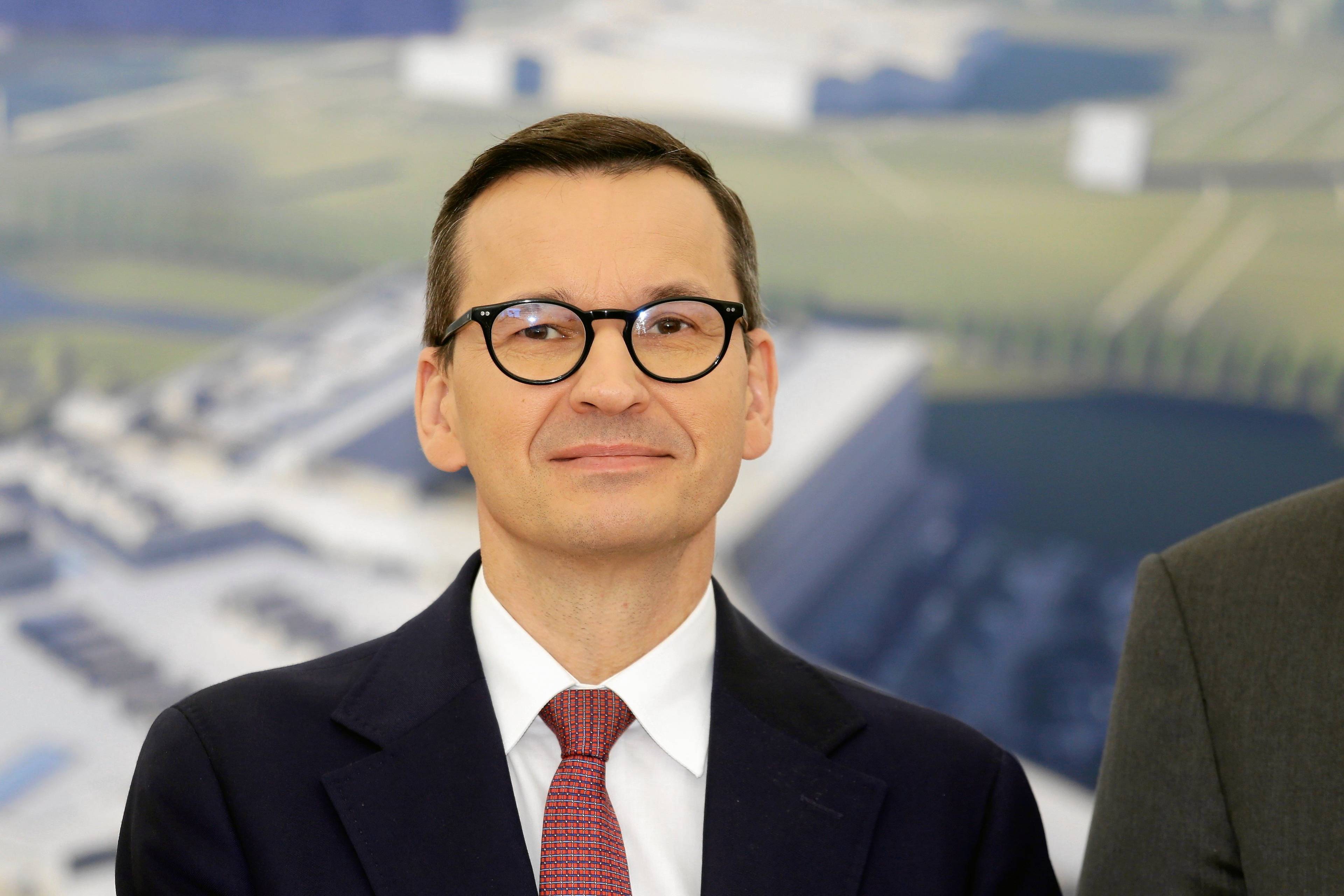 Mężczyzna w okularach i garniturze (premier Mateusz Morawiecki) lekko się uśmiecha, stoli na tle zdjęcia projektu szpitala