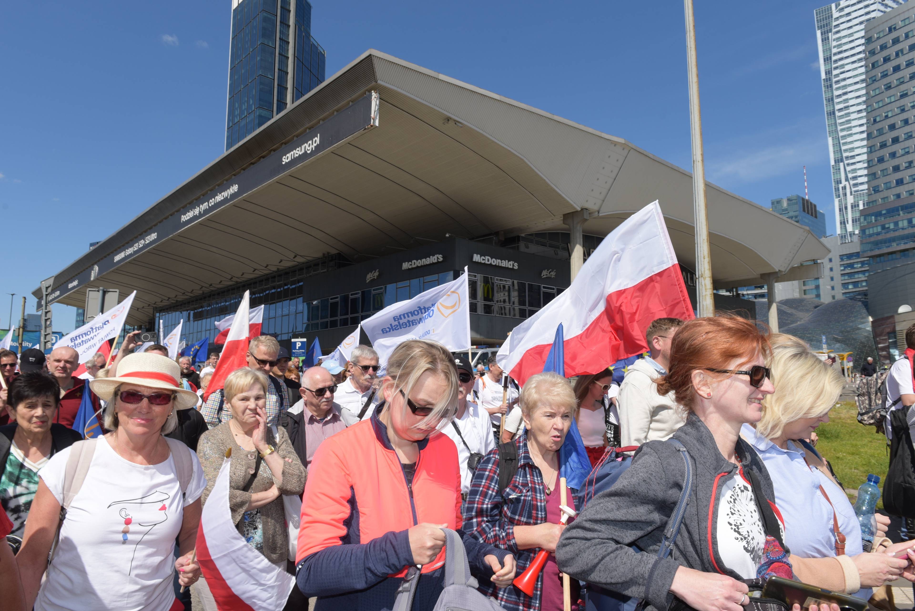 Ludzie z biało-czerwonymi flagami przed Dworcem Centralnym w Warszawie, w tle szklane biurowce