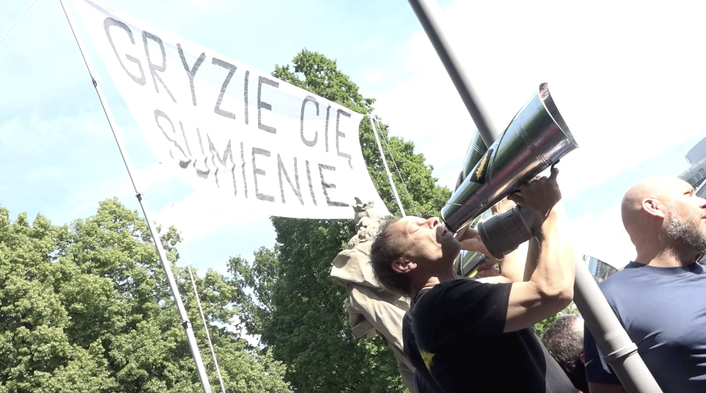 Demonstranci trzymają transparent z napisem „Gryzie cię sumienie” i krzyczą hasła przez metalową tubę. Lotna Brygada Opozycji
