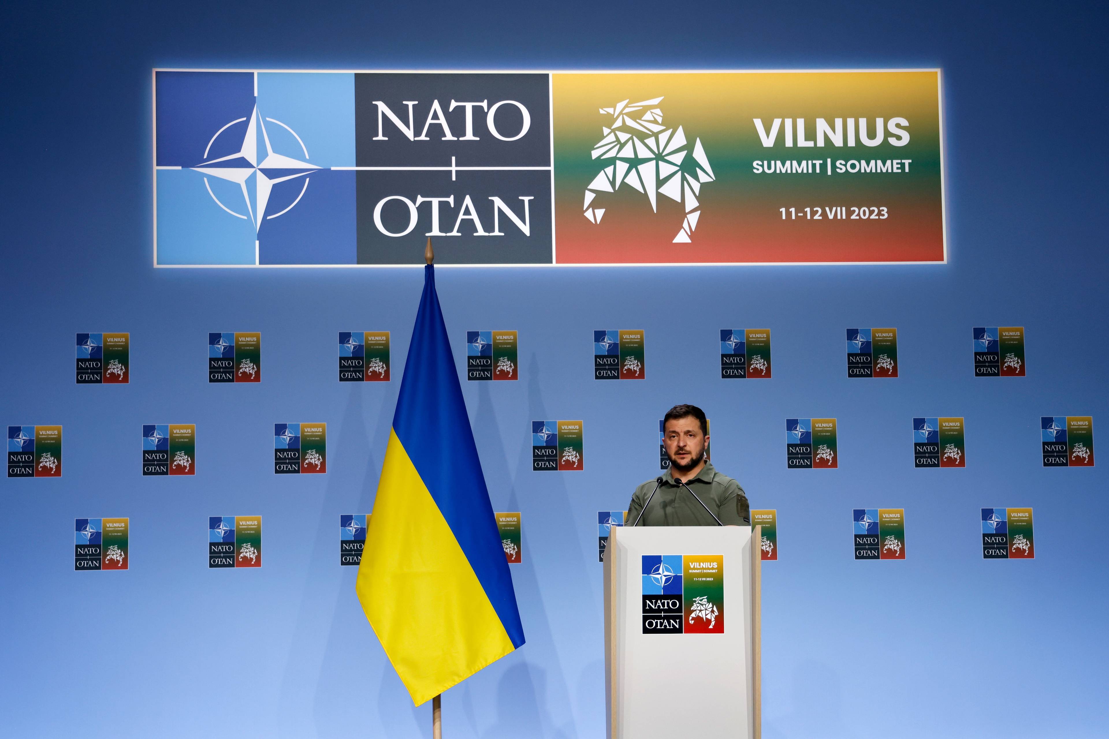 Mężczyzna za mównicą w stroju w kolorze khaki, obok niego flaga Ukrainy, nad nim logo NATO