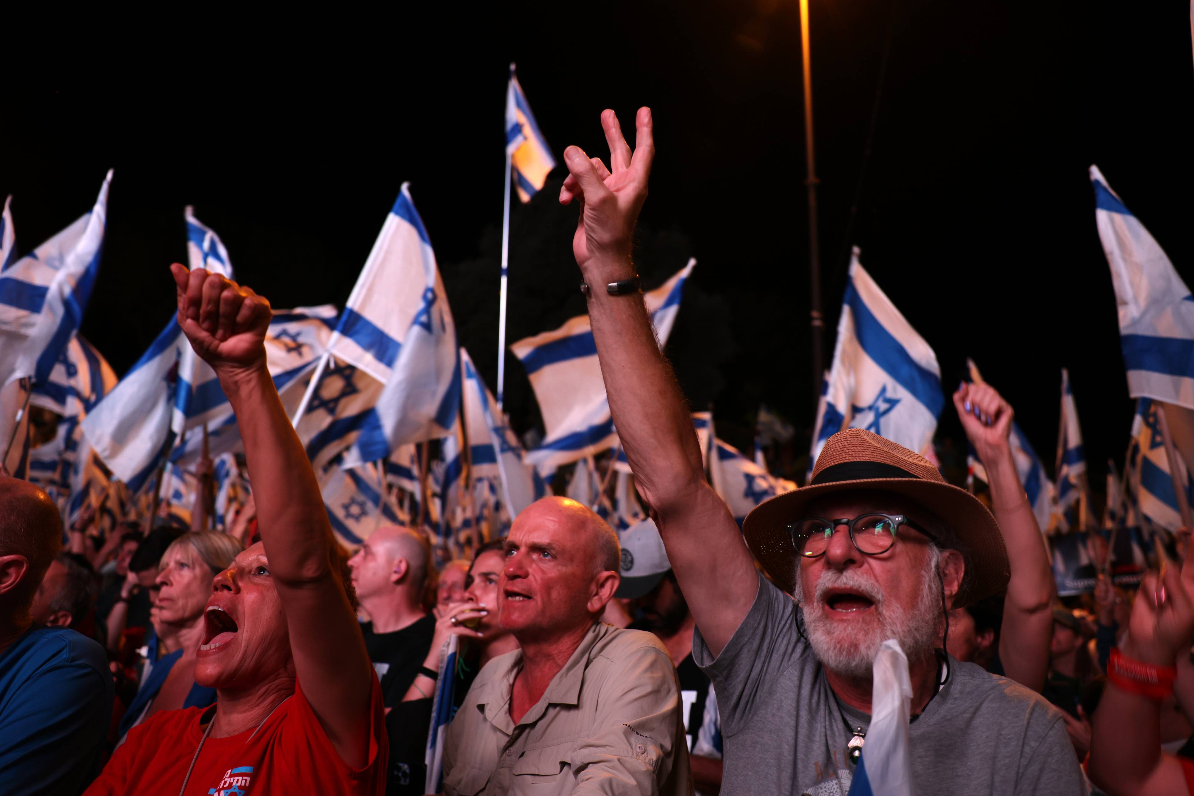 starsi mężczyźni głośno protestujący z podniesionymi w górę rękami, w tle izraelskie flagi