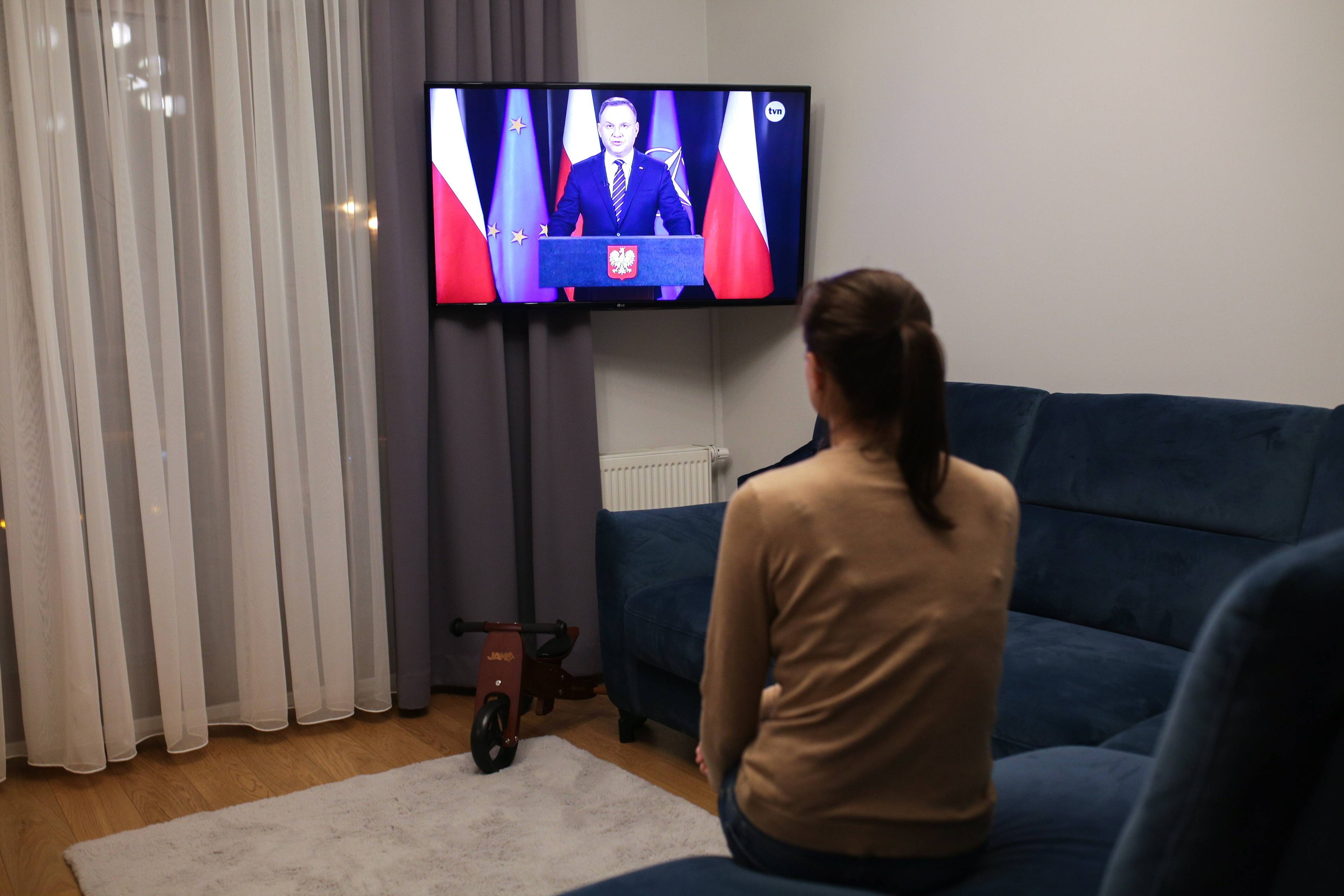 Kobieta siedzi na kanapie, patrzy w telewizor, na którym przemawia prezydent Andrzej Duda