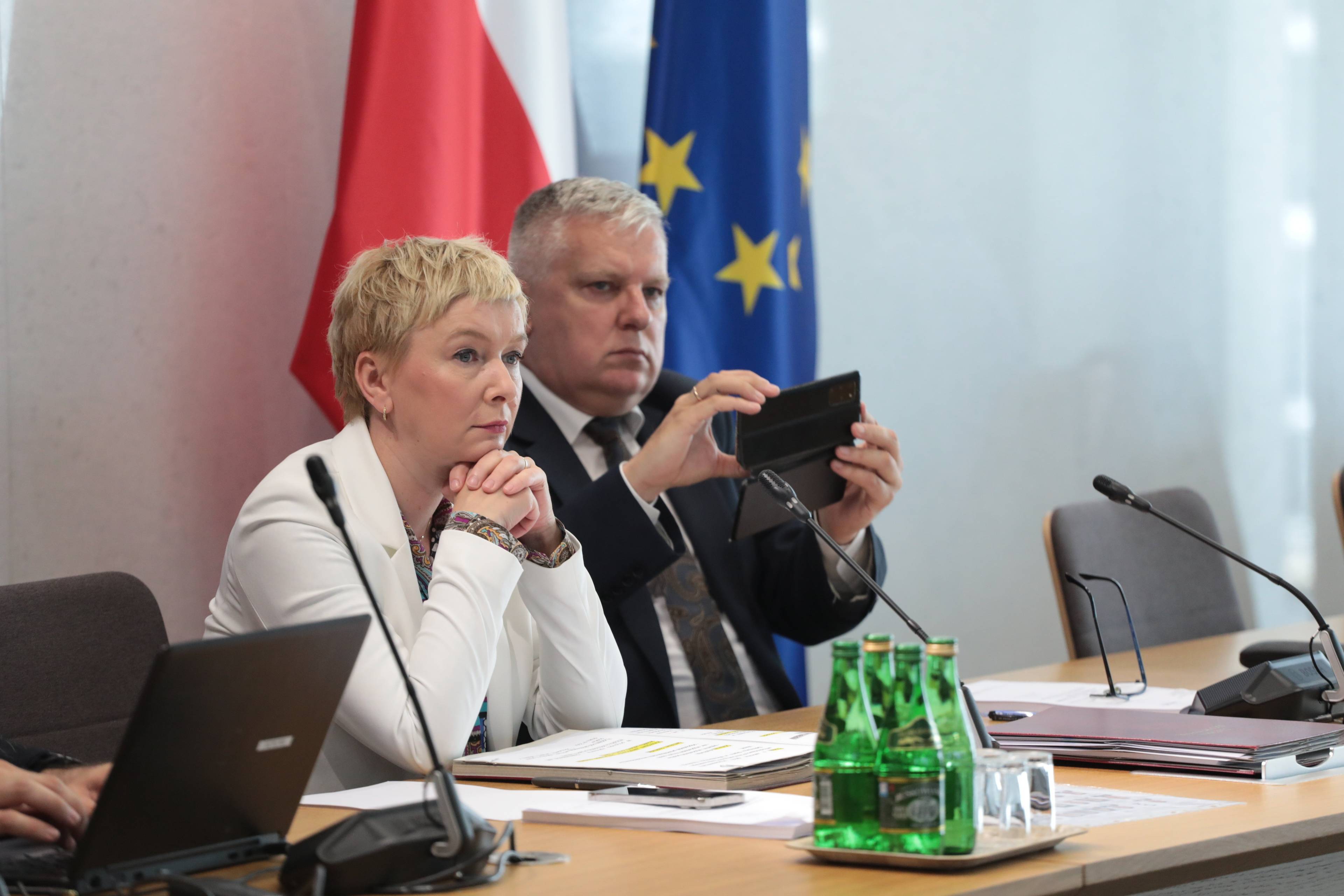Kobieta w białej marynarce razem z mężczyzną w garniturze siedzą przy stole, mężczyzna robi zdjęcie smartfonem, w tle stoją flagi Polski i Unii Europejskiej