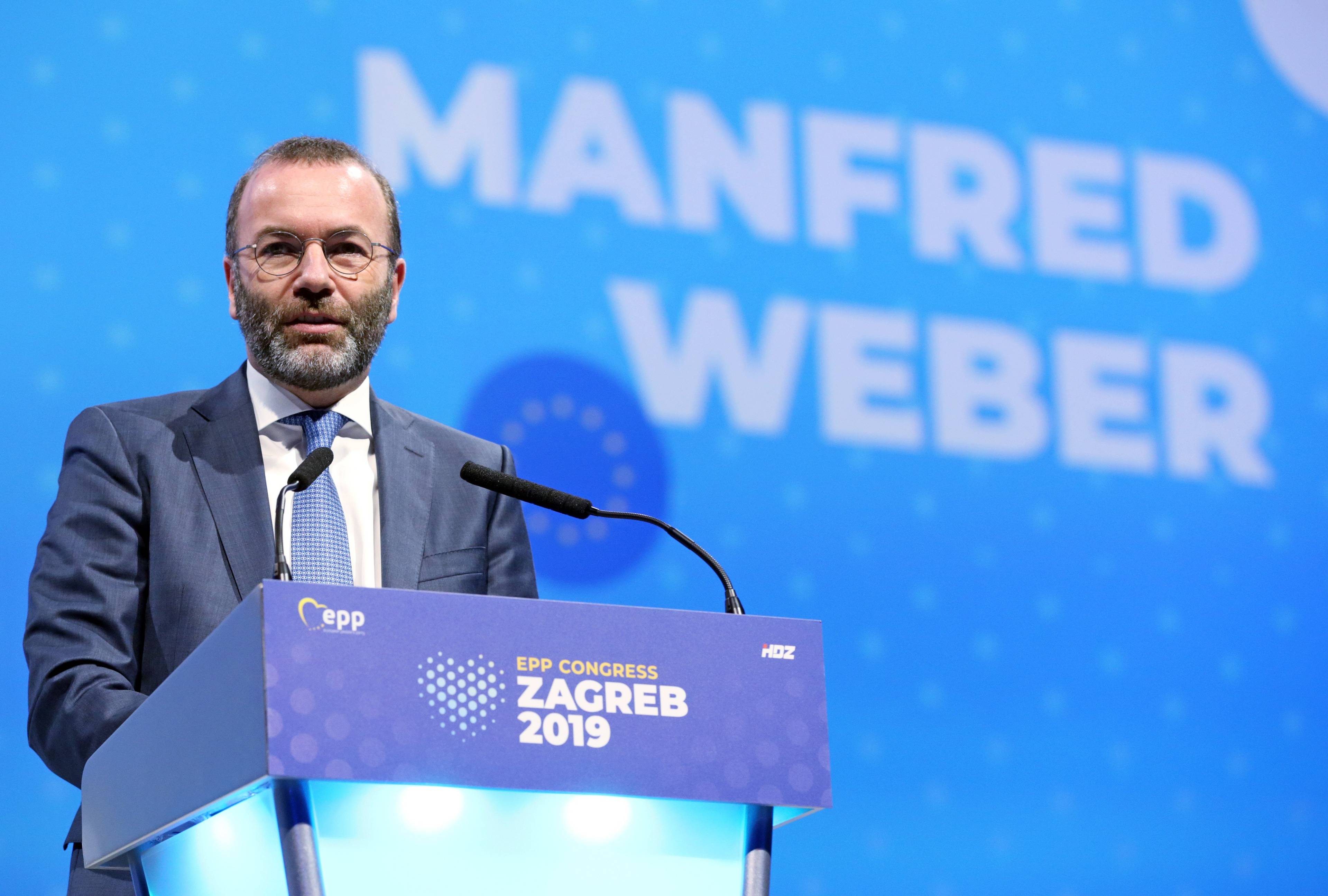 Mężczyzna z krótkim zarostem i w okularach stoi przed mównicą, za nim napis na niebieskim tle o treści: Manfred Weber