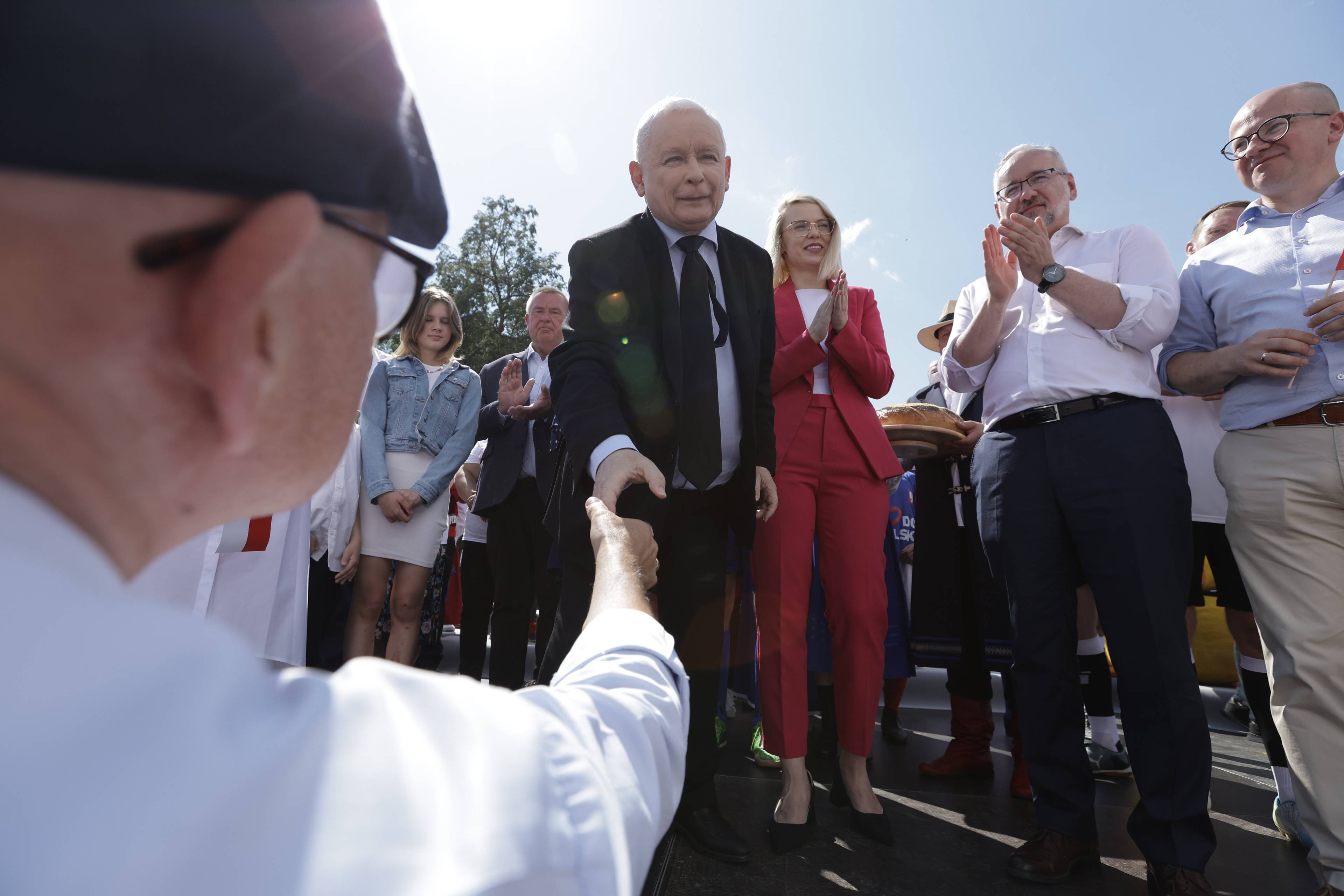 Dwóch starszych mężczyzn podaje sobie rękę, jeden w kaszkiecie i białej koszuli na pierwszym planie, drugi w czarnym garniturze i krawacie (to Jarosław Kaczyński, prezes PiS) na drugim planie