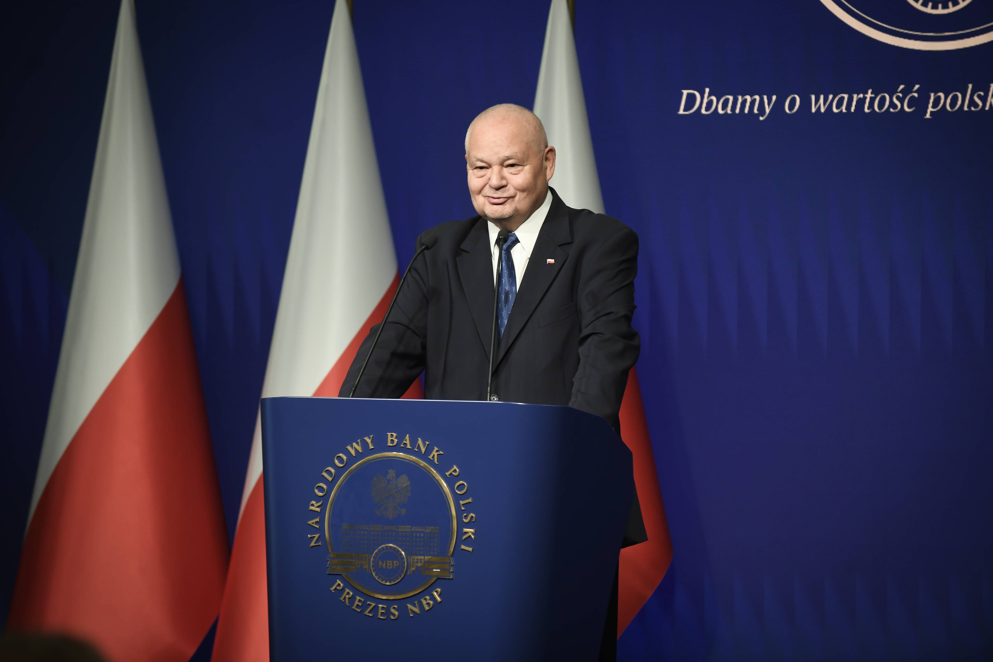 Mężczyzna w garniturze stoi za pulpitem z napisem "Narodowy Bank Polski", w tle stoją trzy polskie flagi
