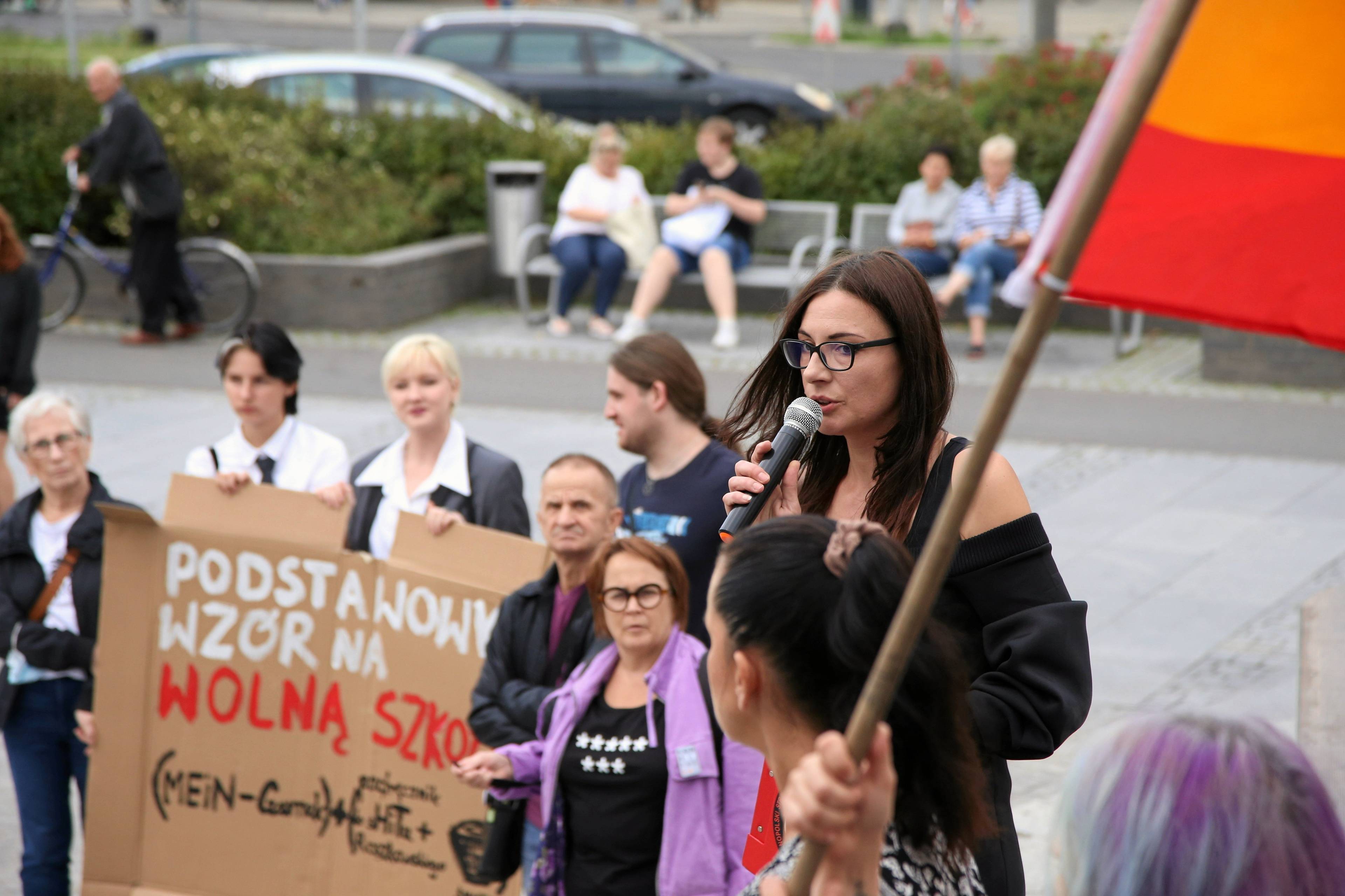 Demonstracja, na pierwszym planie kobieta przemawia przez mikrofon, w tle demonstranci trzymają karton z hasłem Podstawowy wzór na wolną szkołę
