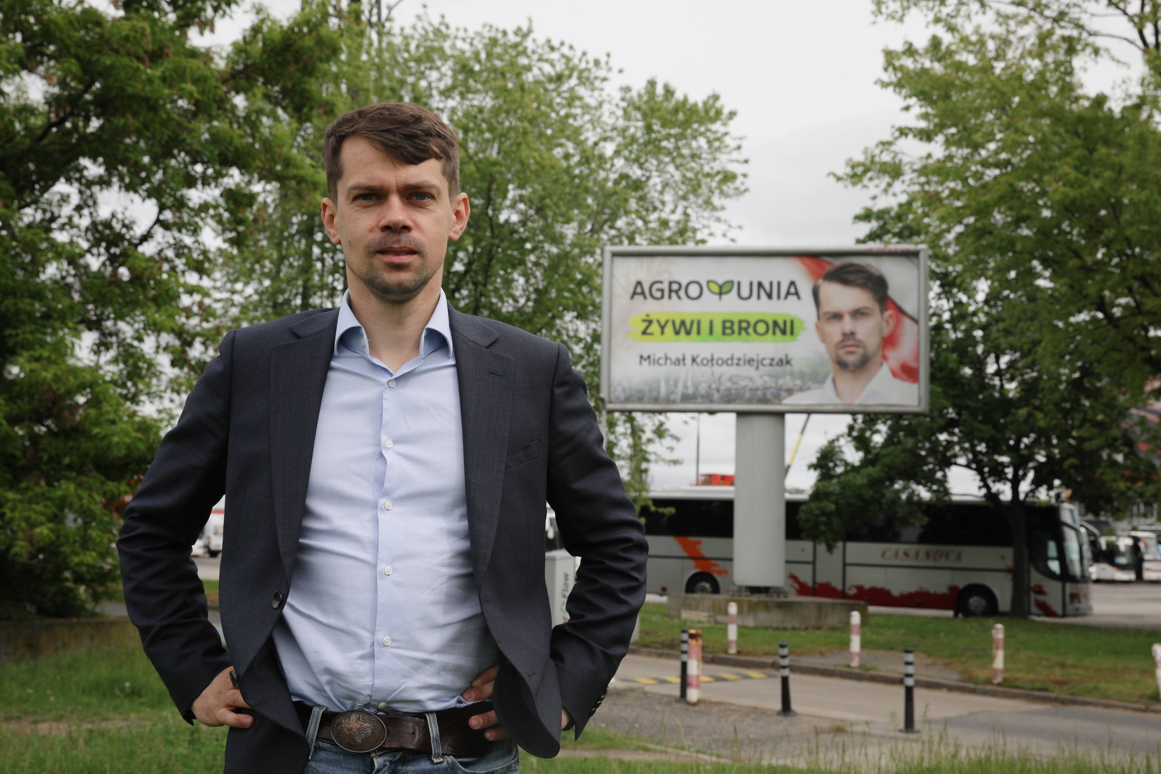 Michał Kołodziejczak – 30-latek z brodą i wąsami stoi podparty pod boki, w tle plakat Agrounii z jego twarzą