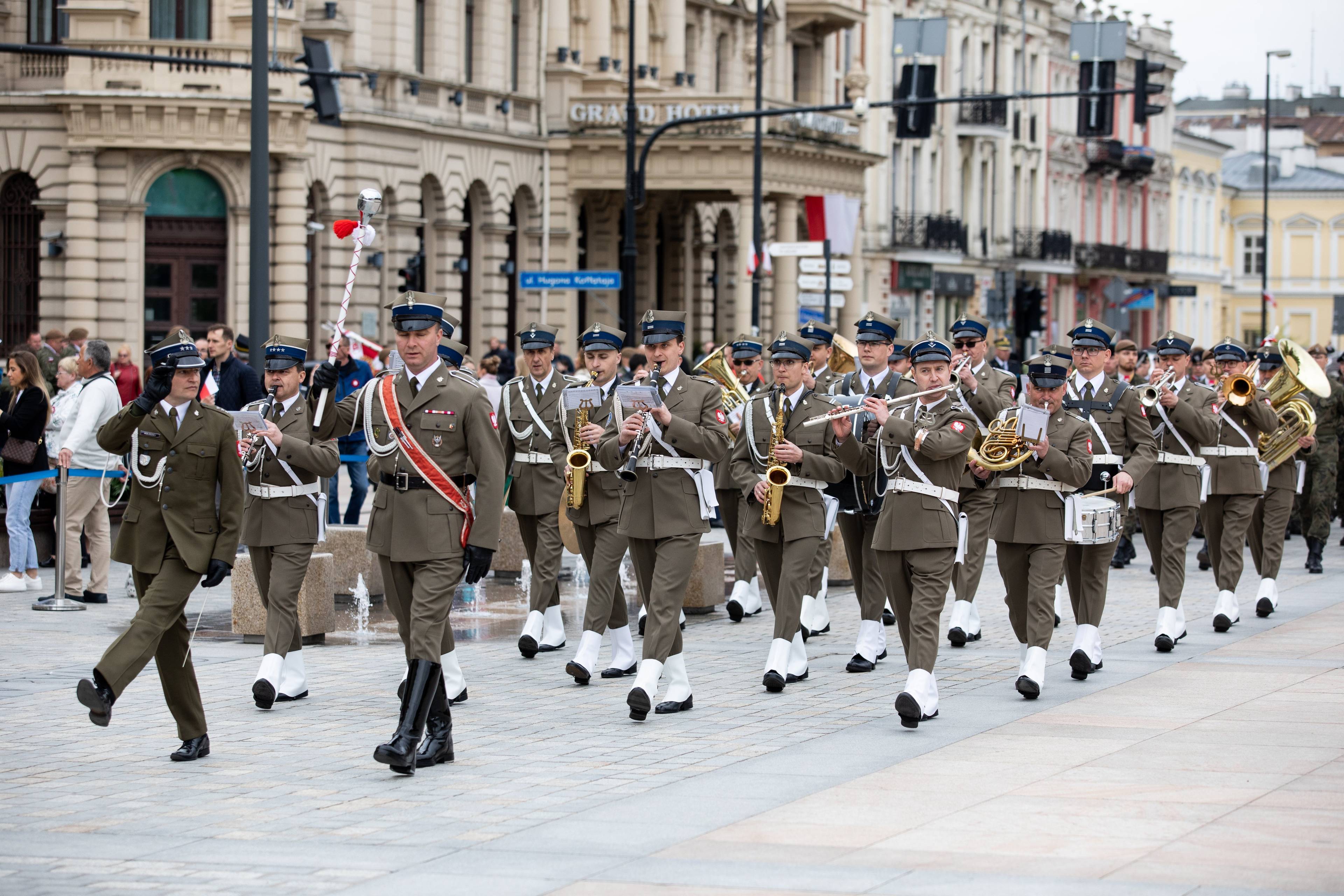 orkiestra wojskowa w stroju galowym maszeruje przez ulicę z XIX-wiecznymi budynkami