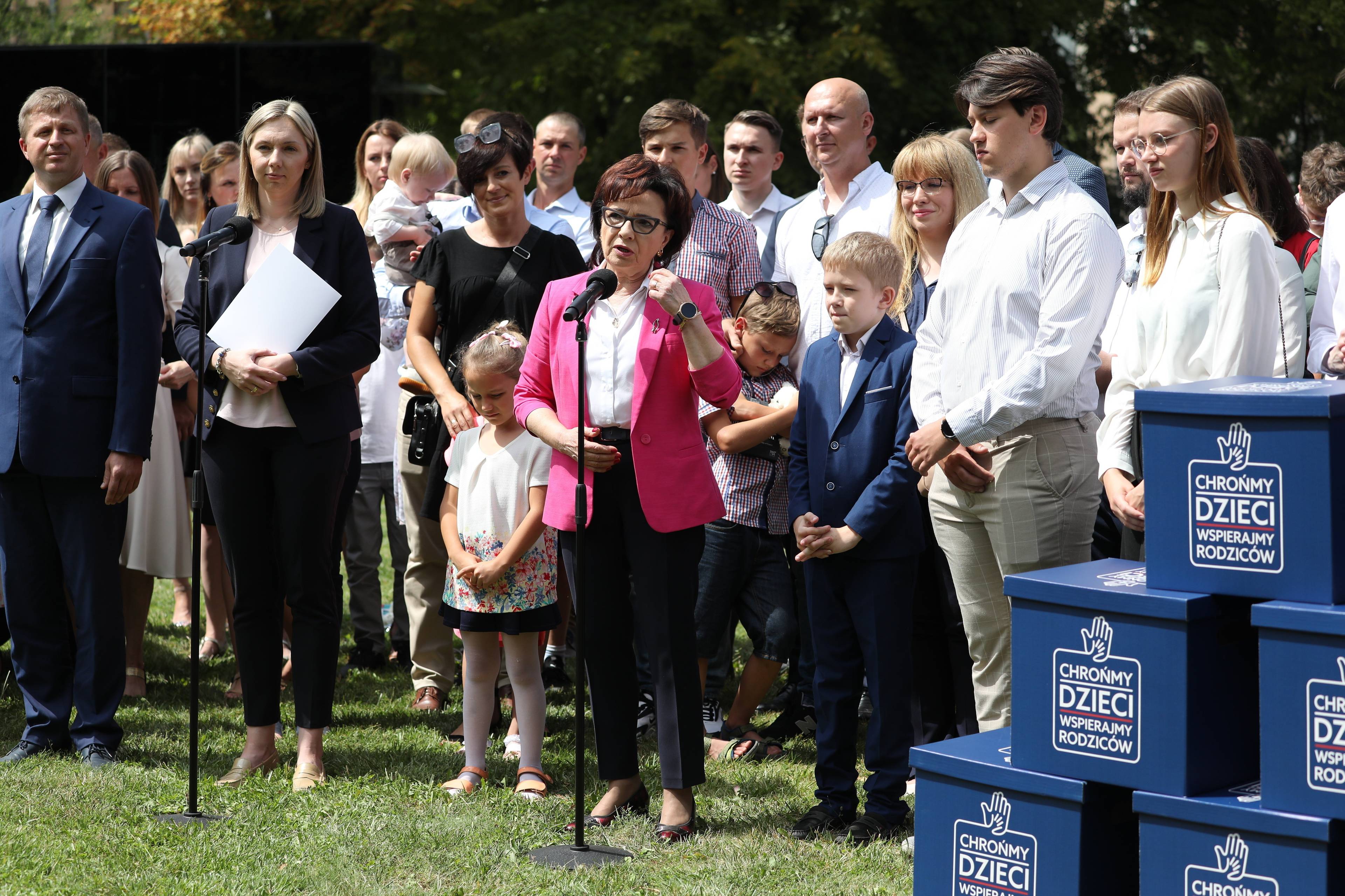 Grupa dorosłych i dzieci stoi na świeżym powietrzu, na pierwszym planie brunetka w purpurowym żakiecie i okularach przed mikrofonem - marszałek Witek