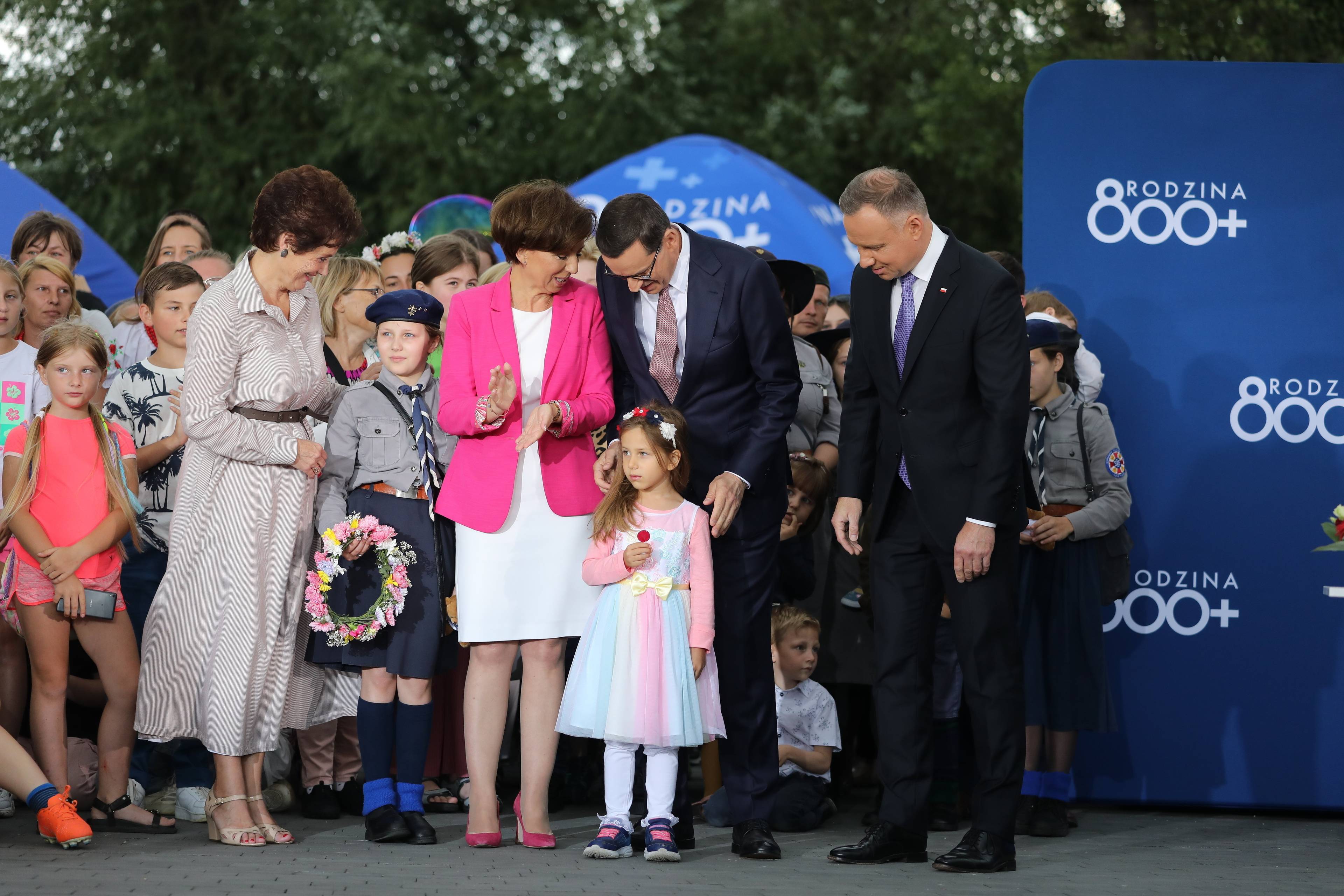 Grupa ludzi na scenie z małą dziewczynką" Ministra Maląg nachyla się do premiera Morawieckiego, na co patrzy prezydent D