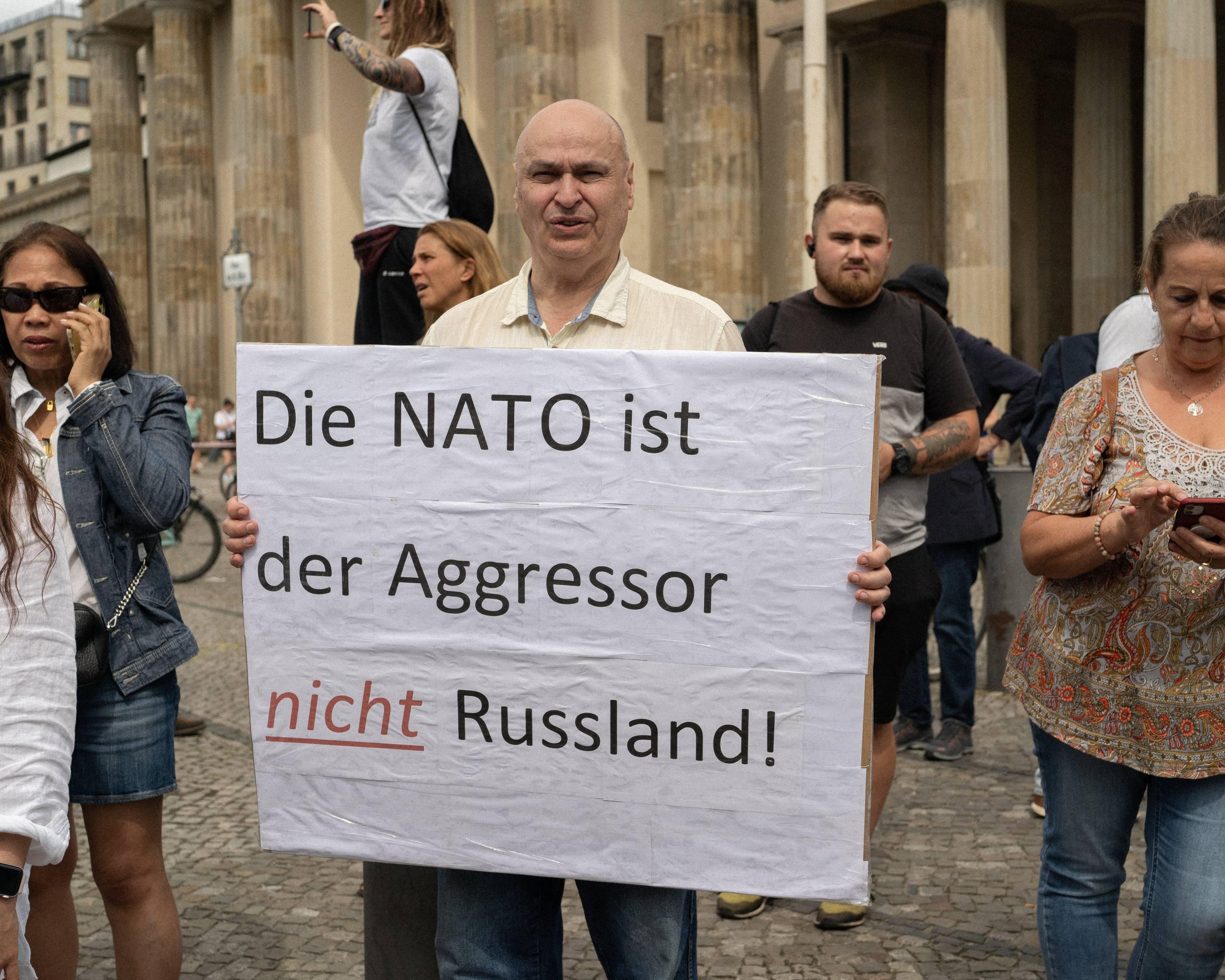 scena z demonstracji, mężczyzna w białej koszuli trzyma transparent z napisem po niemiecku :NATO jest agresorem, nie Rosja