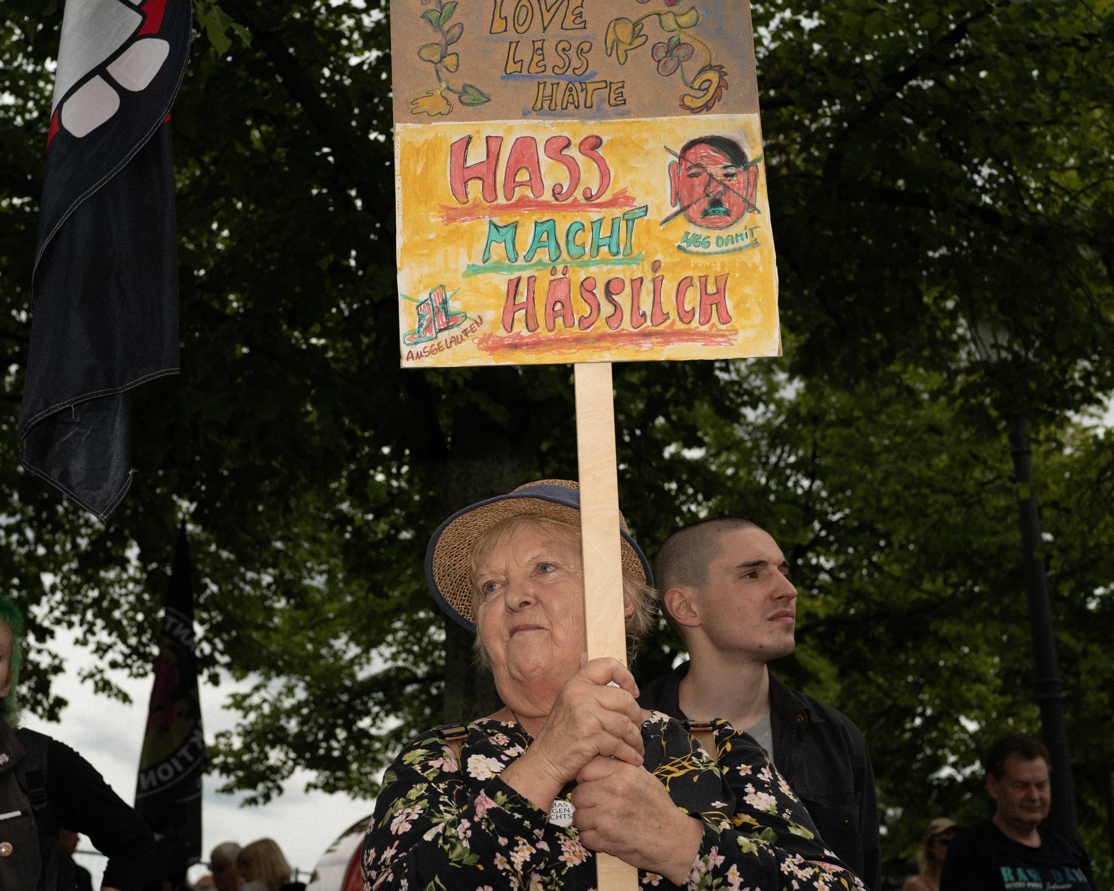 Starsza pani w słomkowym kapeluszu trzyma transparent z hasłem po niemiecku i przekreśloną karykaturą Hitlera