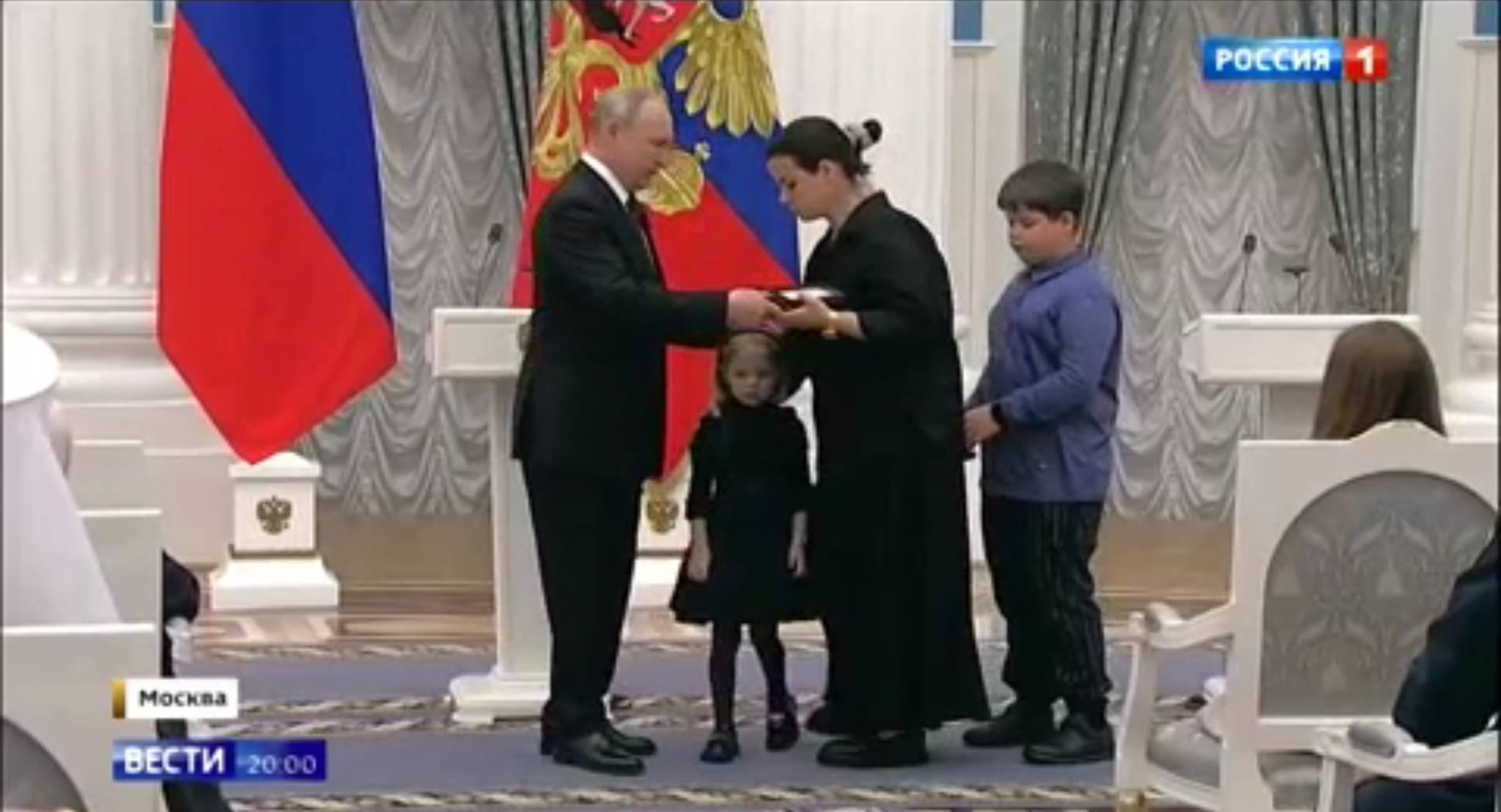 Kobieta w czerni z dwójka małych dzieci odbiera dyplom z rąk Putina
