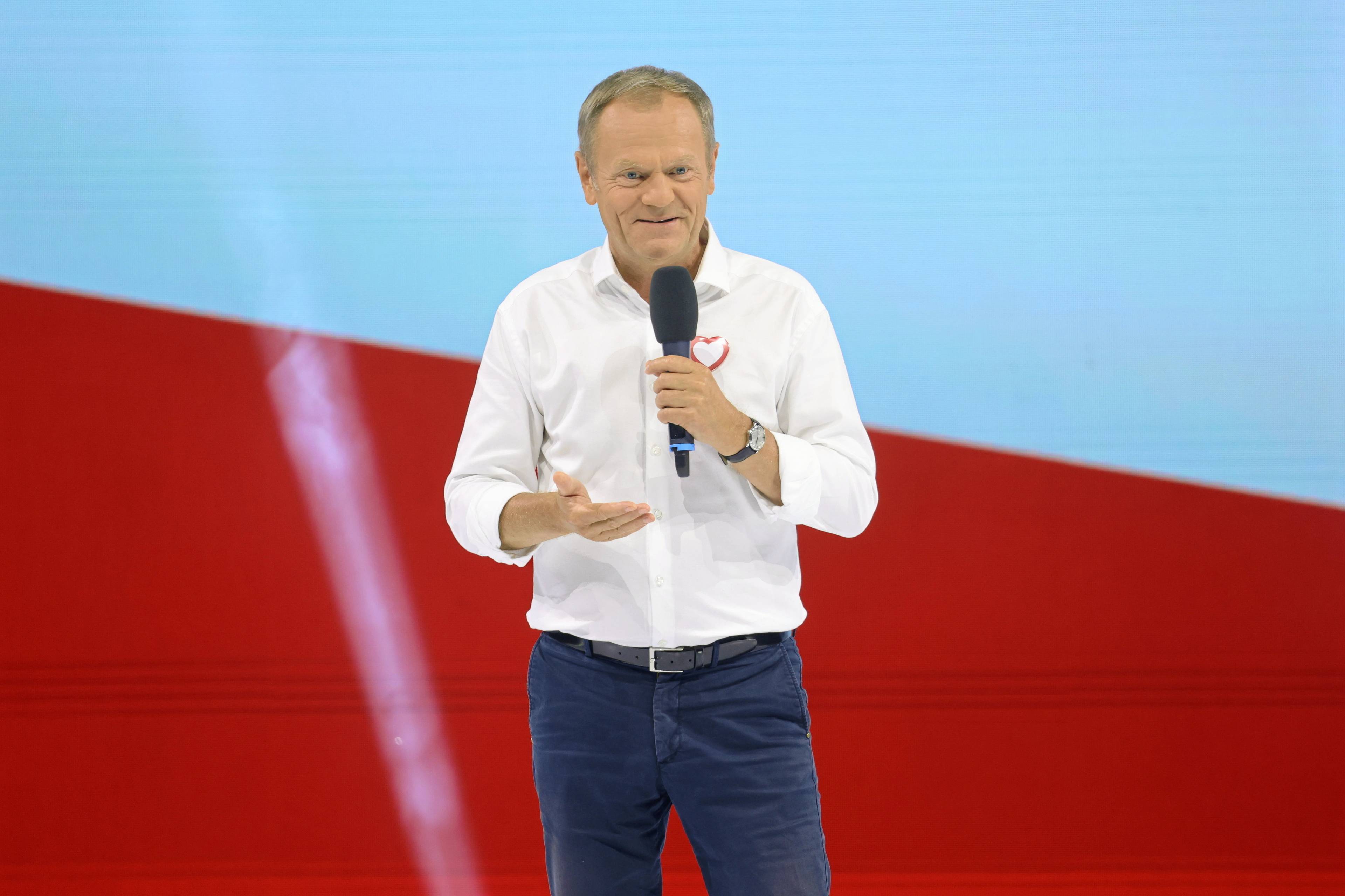 Donald Tusk przemawia w białej koszuli na tle polskiej flagi