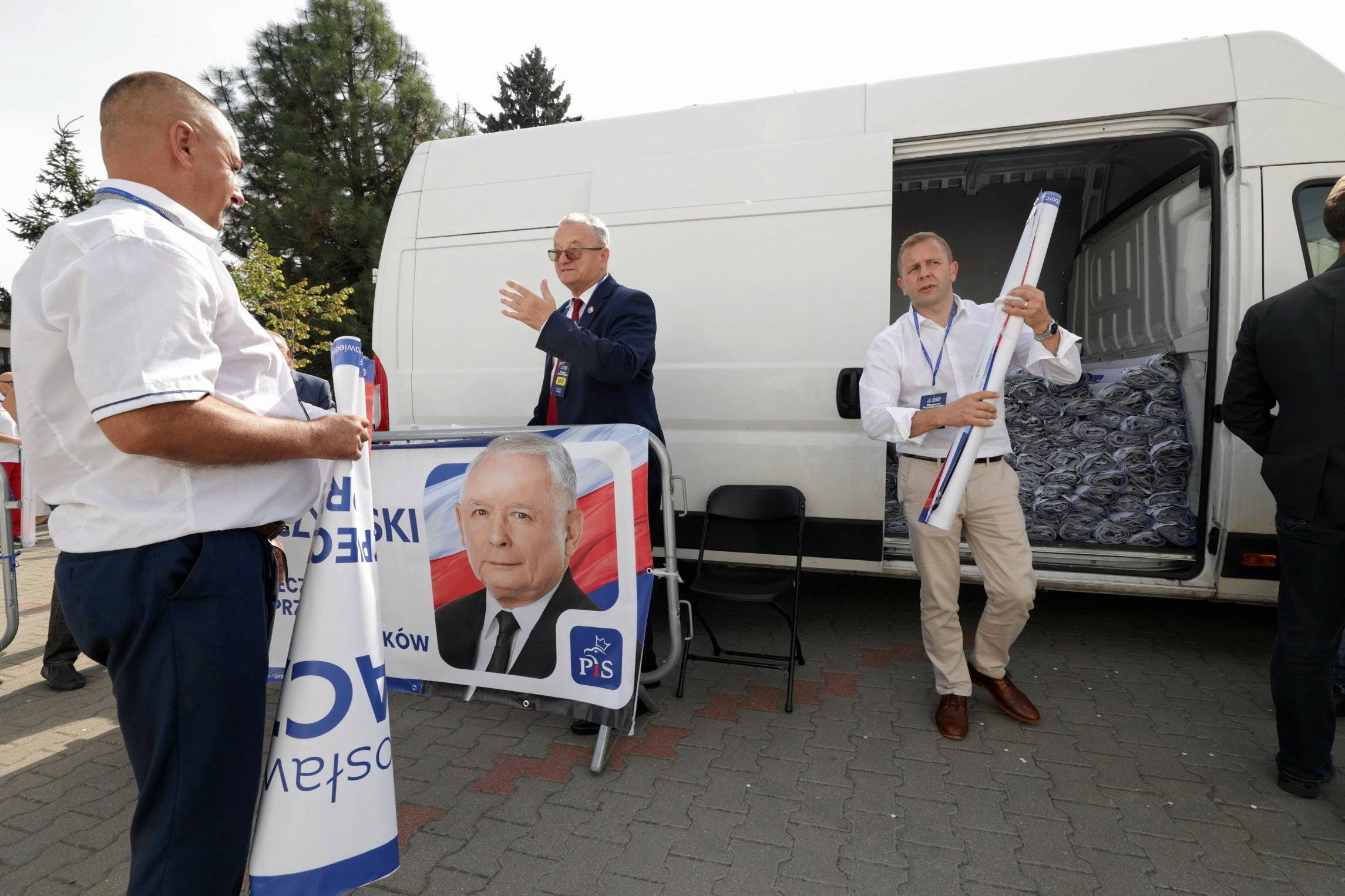 trzech mężczyzn wypakowuje z białego minvana baner z Jarosławem Kaczyńskim i inne materiały na kampanię wyborczą