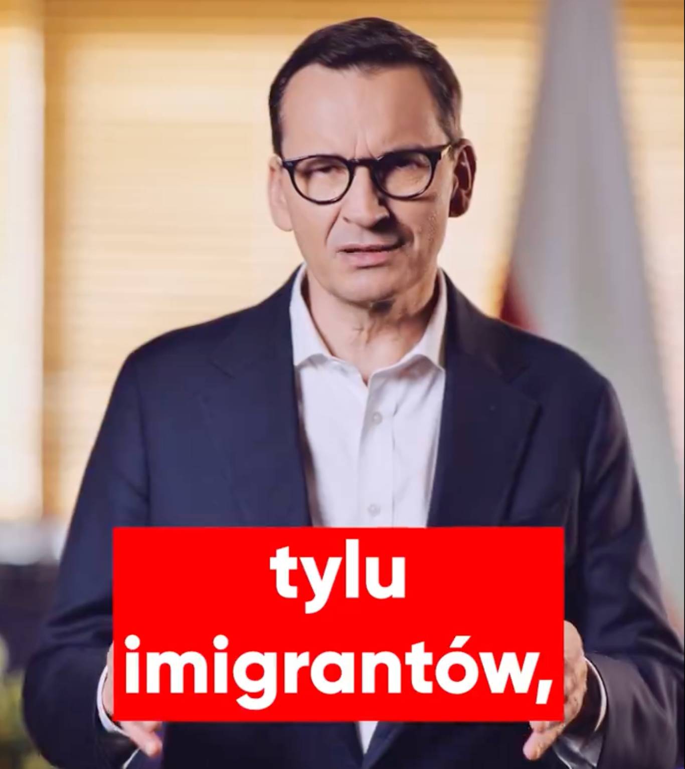 Premier Mateusz Morawiecki przemawia na filmie wideo, u dołu ekranu na czerwonym tle słowa „tylu migrantów”