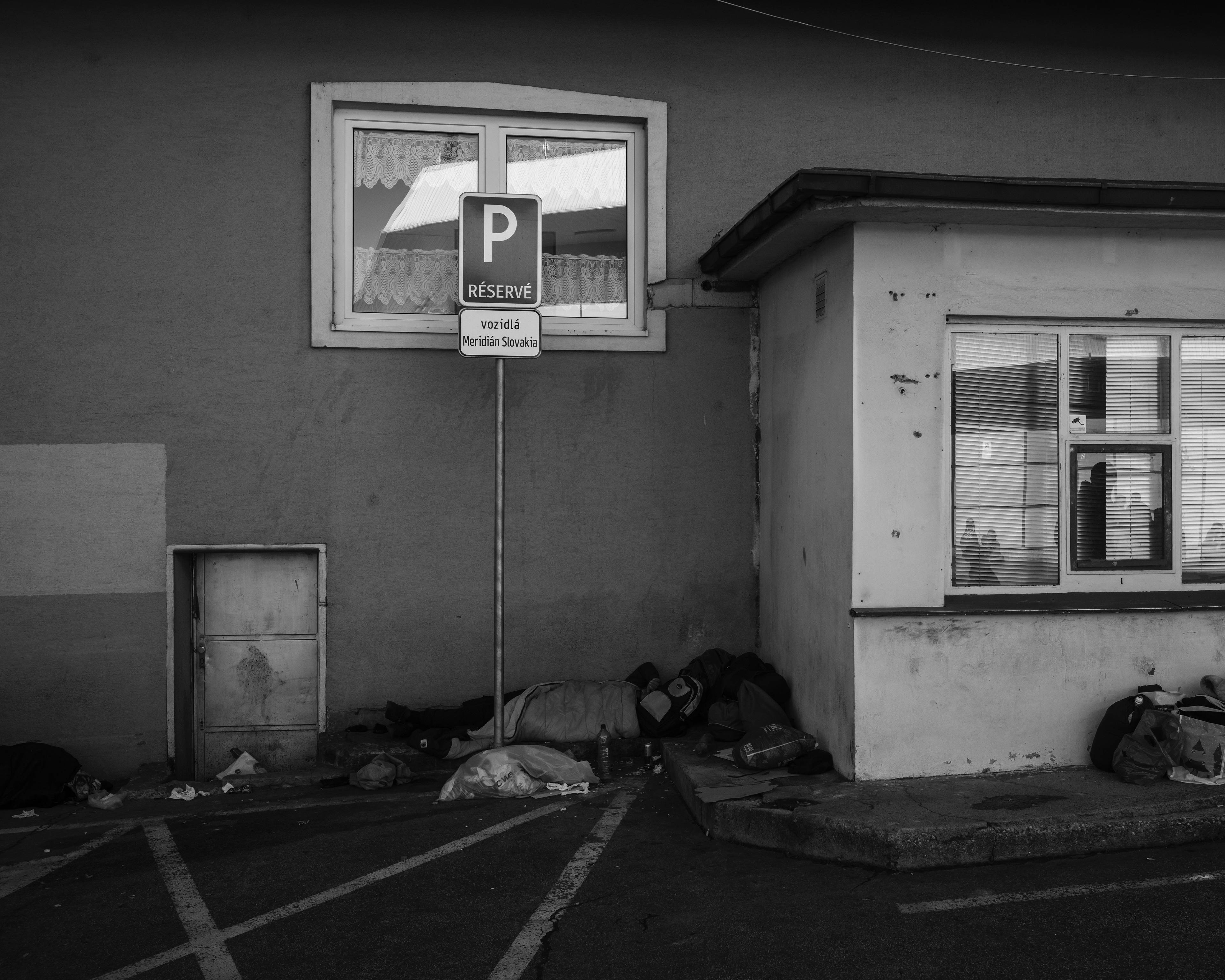 pusty parking przed budynkiem, pod ścianą ktoś leży owinięty w śpiwór, zdjęcie czarno-białe