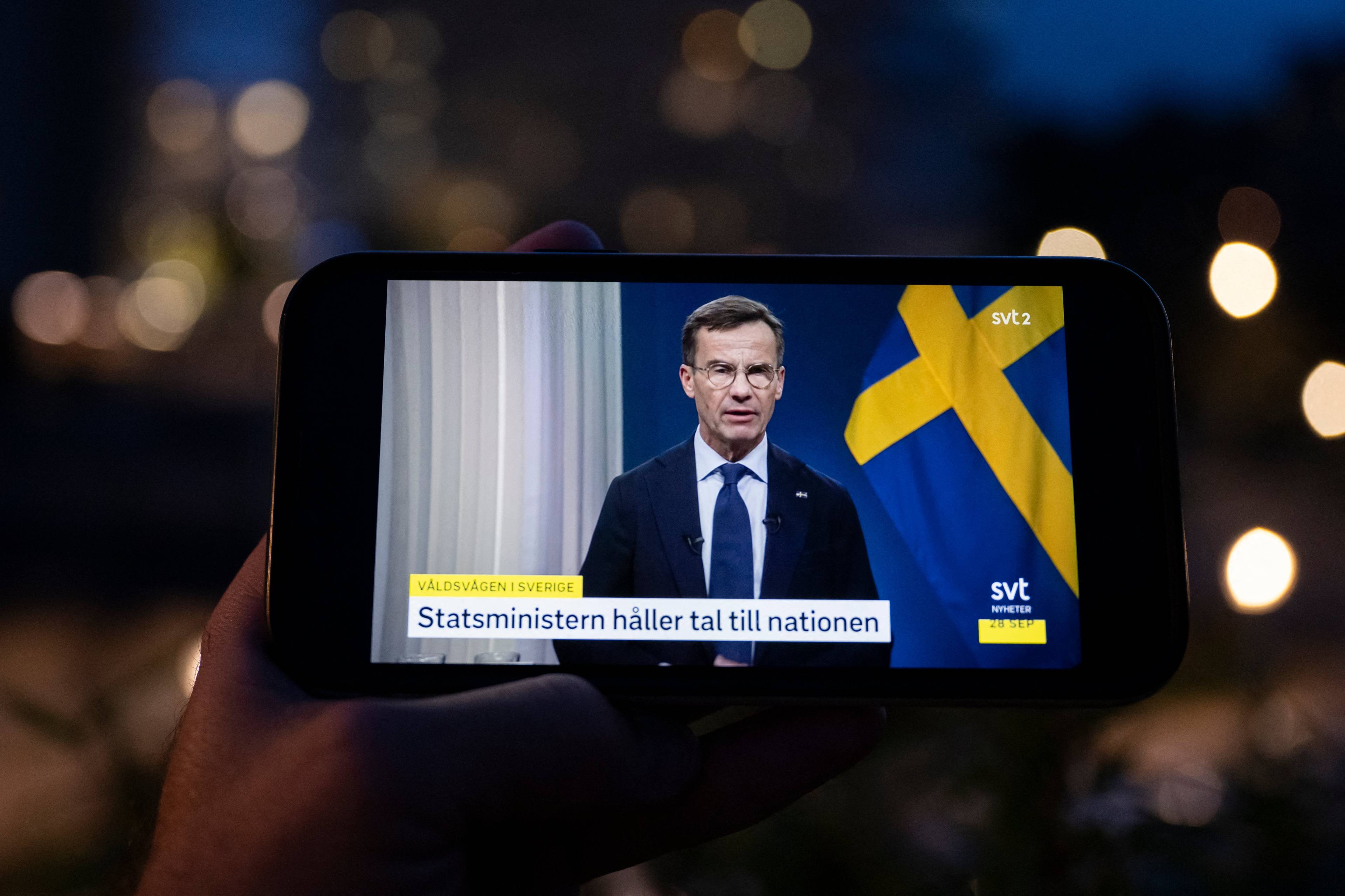 Na ekranie telefonu widać męzczyznę w garniturze, który przemawia przy szwedzkiej fladze. Napis po szwedzku